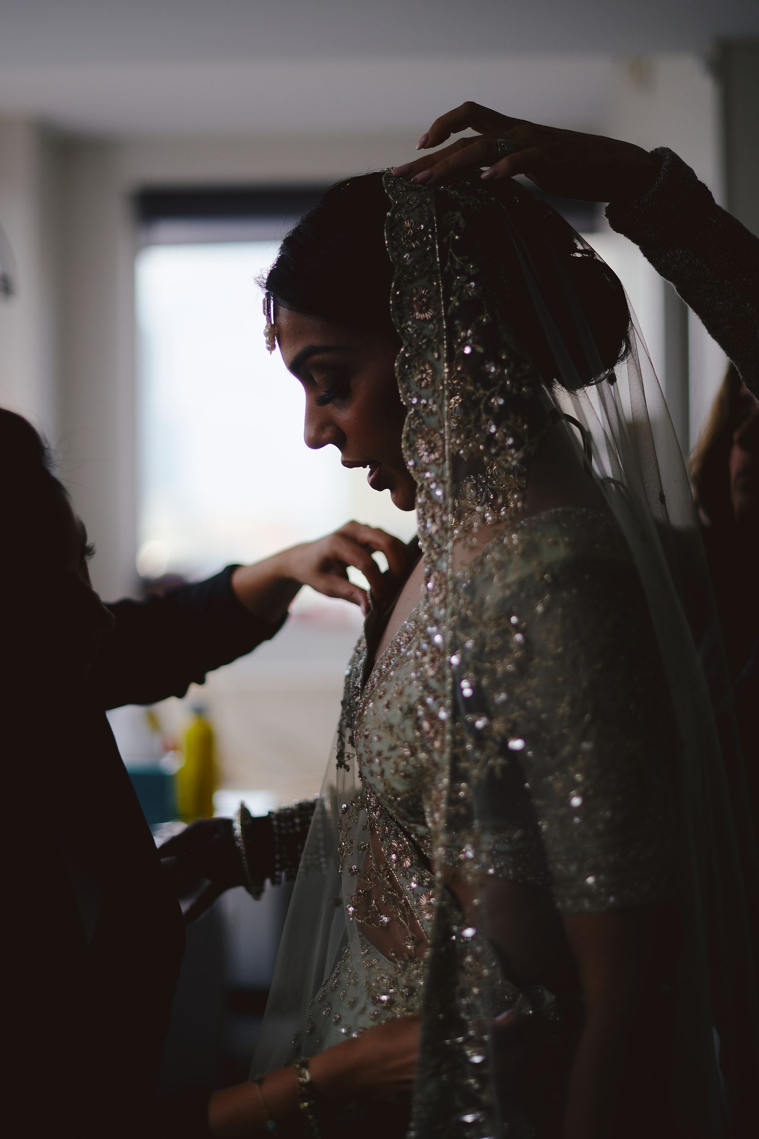 A silhouette of a bride getting ready, with sparkly details visible on her veil and dress in a backlit setting.