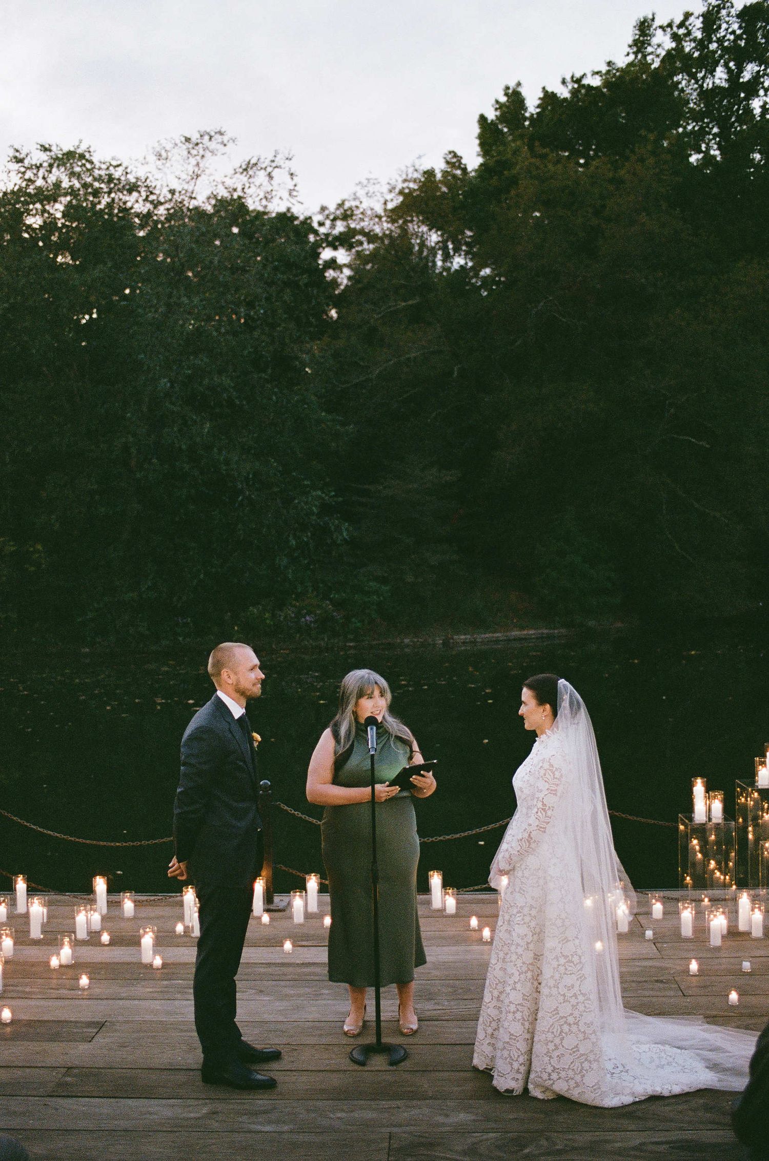 A romantic outdoor wedding ceremony on a wooden deck illuminated by glowing candles at dusk with trees in the background.