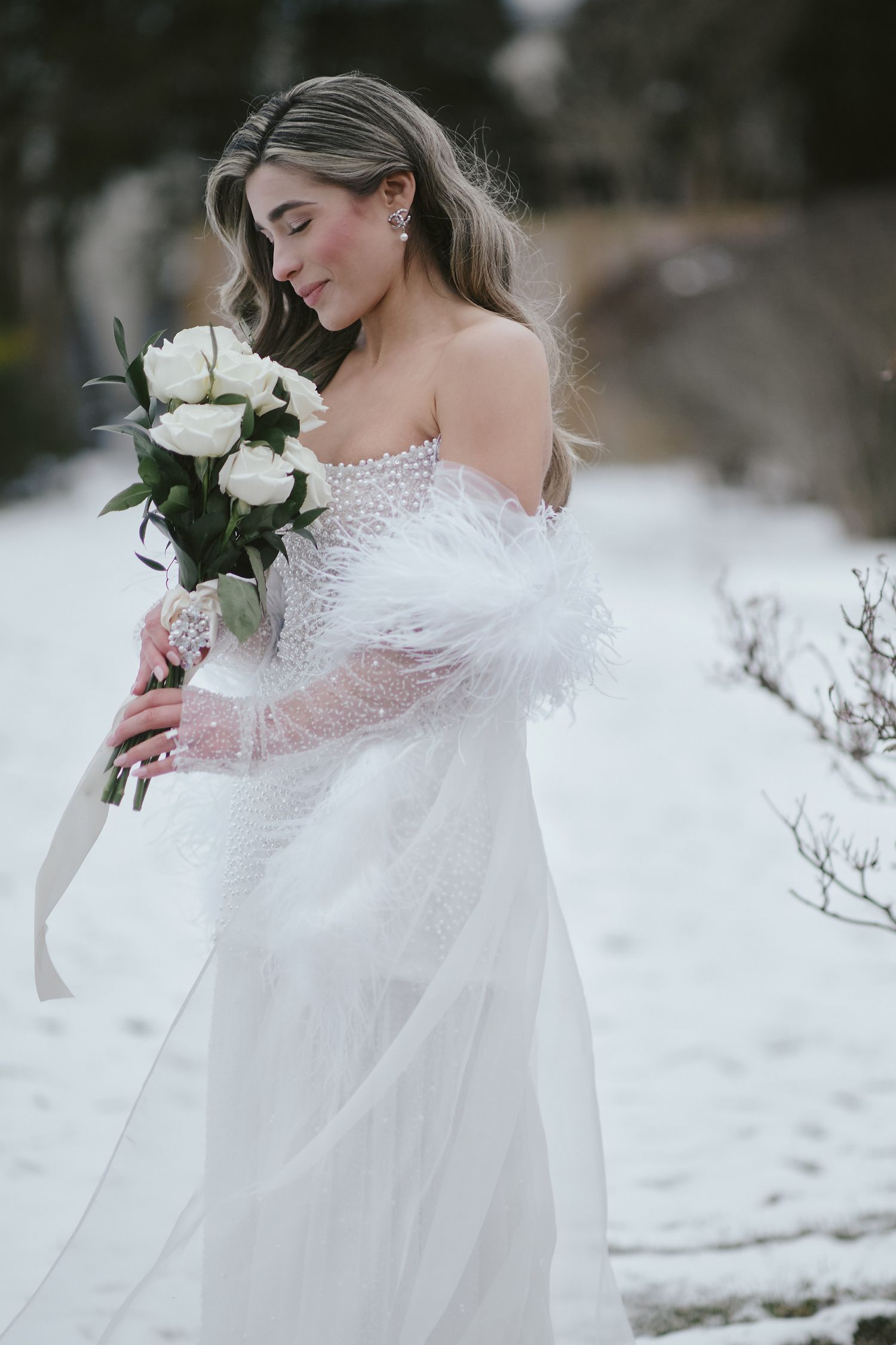 A bride in an ethereal white off-shoulder lace gown holds white roses while standing in a snowy winter landscape.