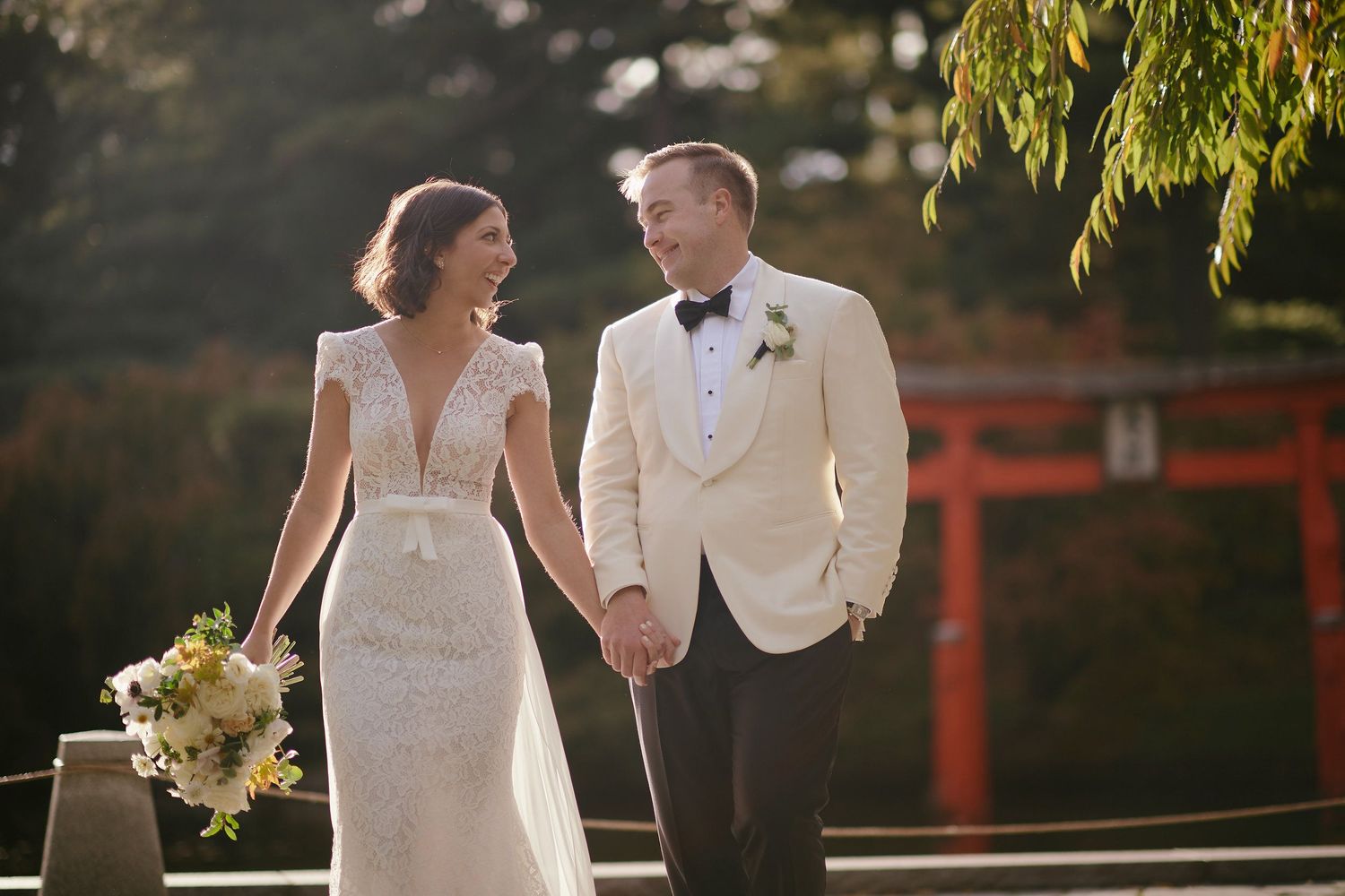 A newlywed couple holds hands while walking together outdoors in formal wedding attire during golden hour.