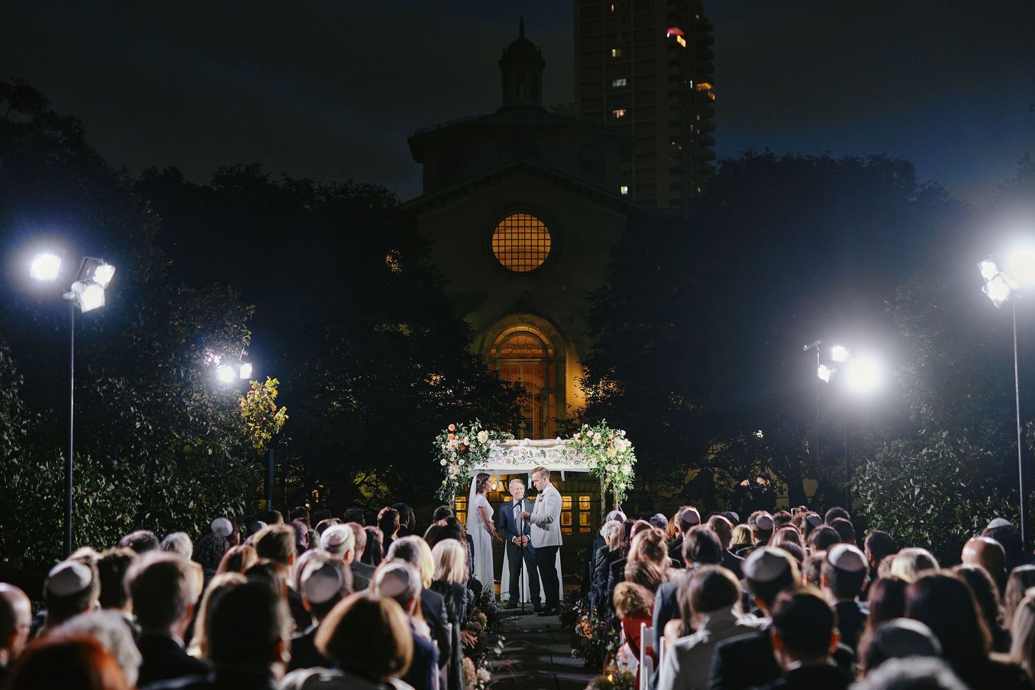 A nighttime outdoor wedding ceremony in front of a brick church illuminated by bright spotlights with a large crowd attending.