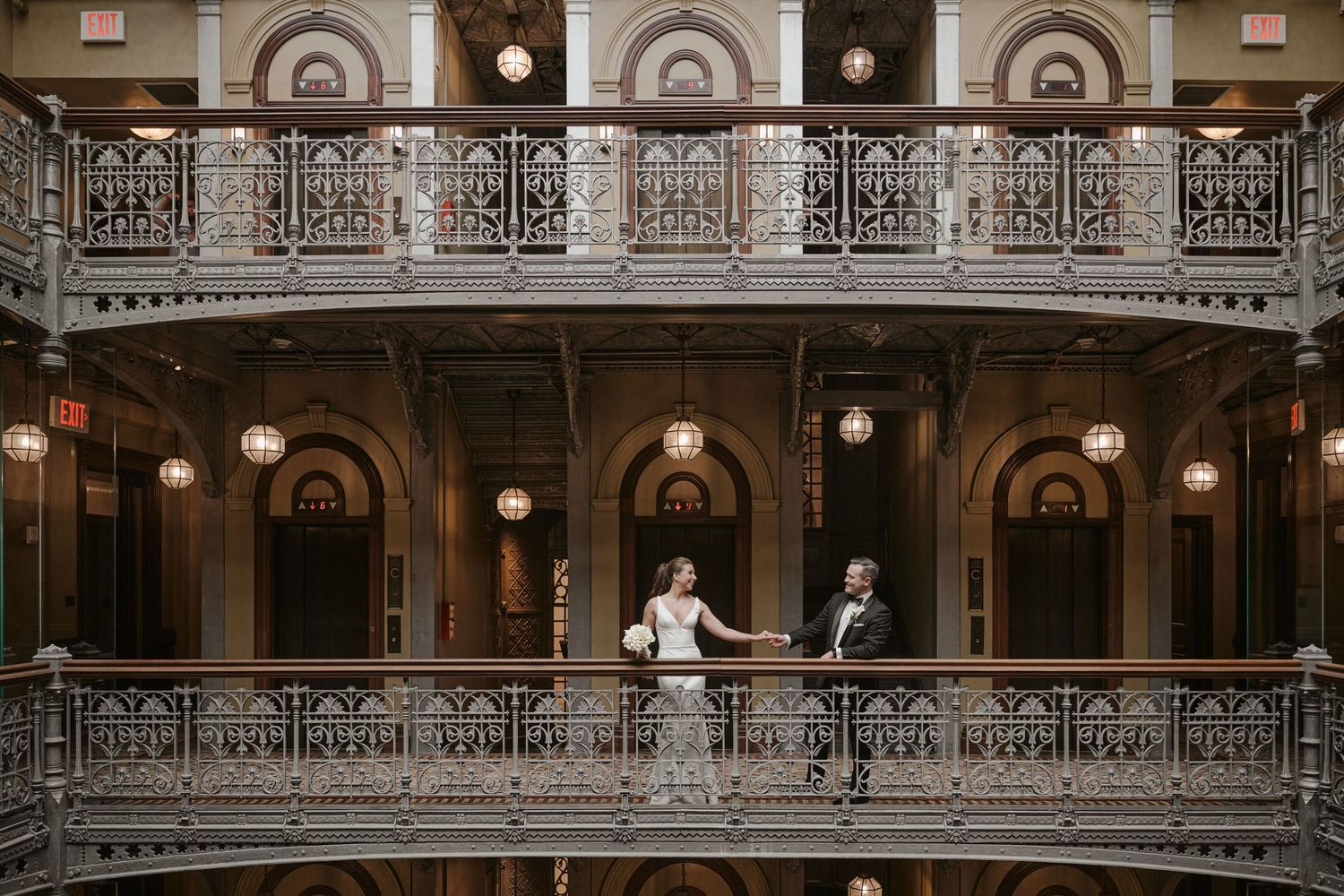 Bride and groom pose on ornate iron balcony of elegant historic building with arched windows and warm lighting.