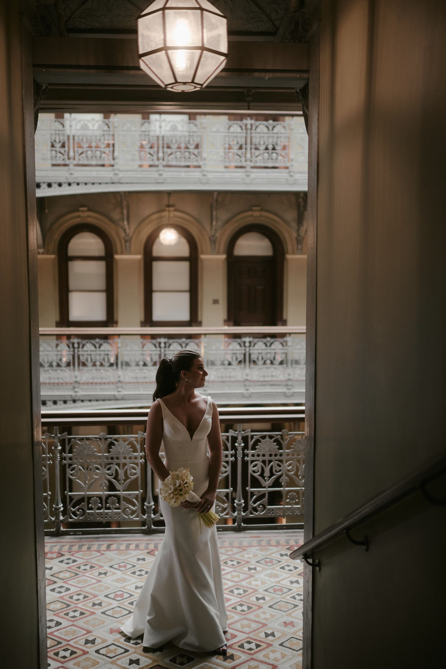 A person in an elegant white wedding dress stands in a vintage architectural building with ornate railings and arched windows.
