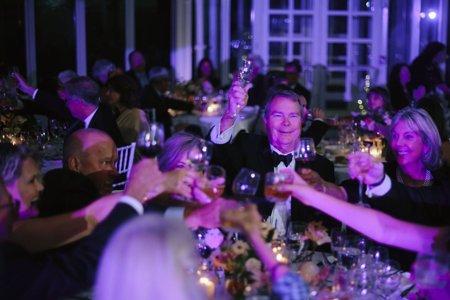 Party guests raise their wine glasses in a toast during an elegant evening event with purple mood lighting.