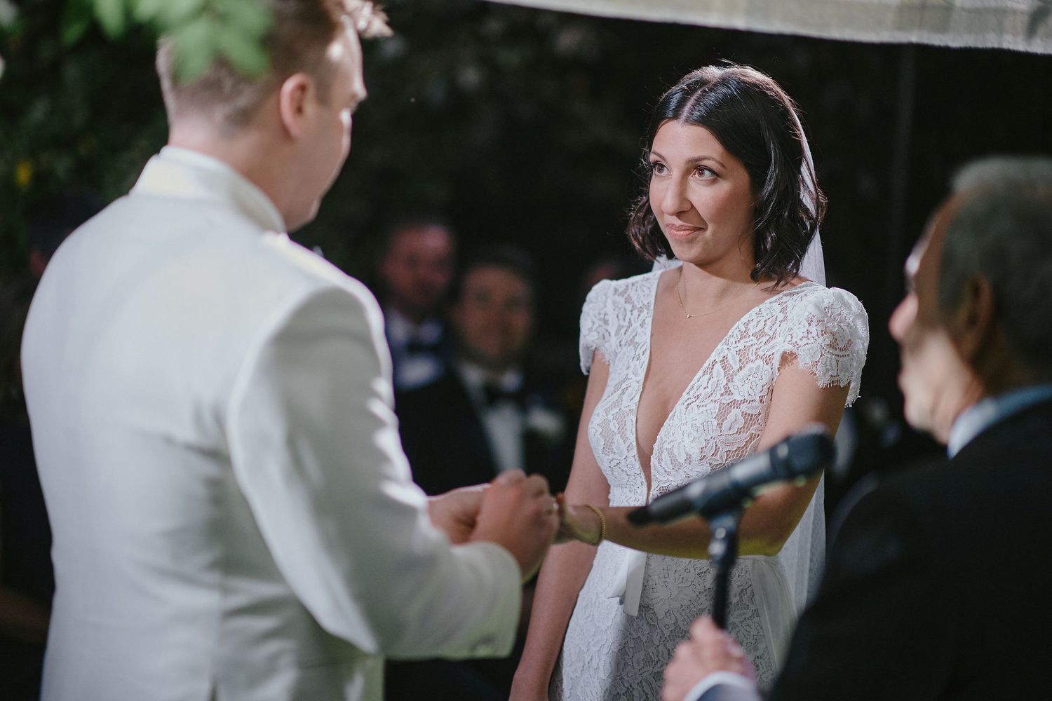 Couple exchanges wedding rings during outdoor ceremony with guests in background.