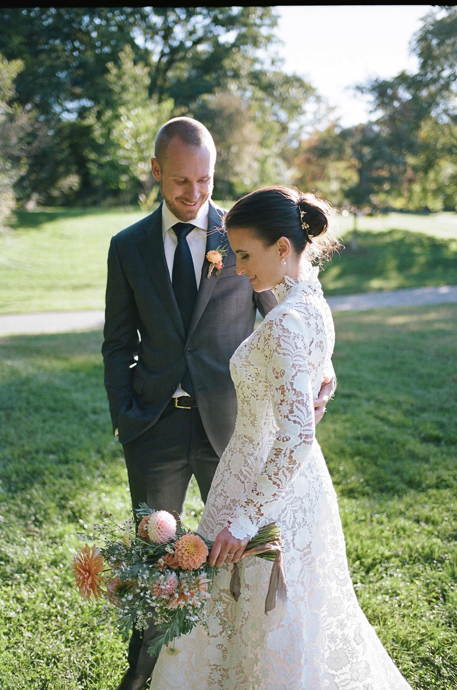 Couple stands together outdoors in wedding attire on a sunny day with lush green lawn and trees in background.