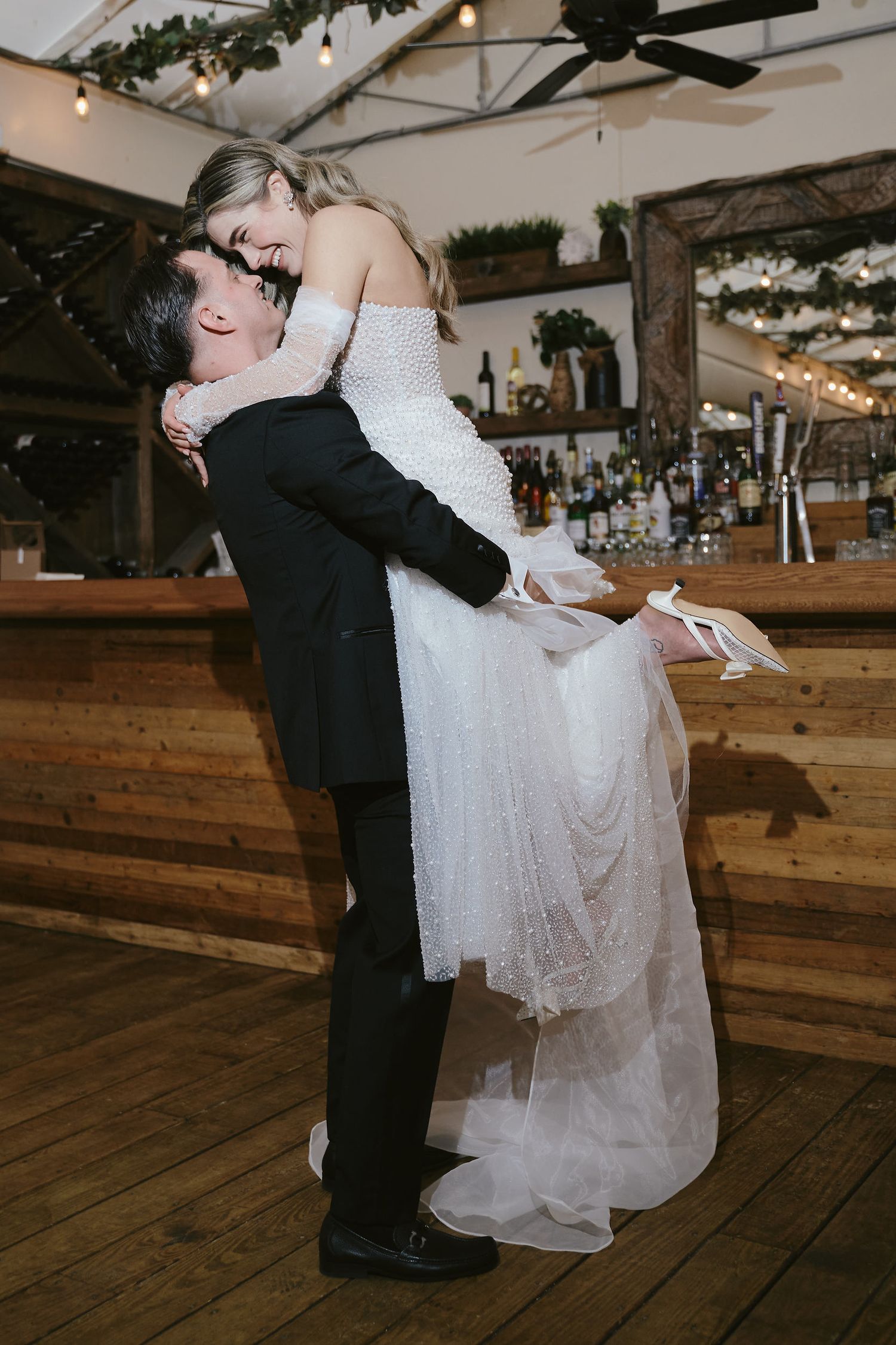 A couple shares a romantic dip kiss at their rustic barn wedding reception next to a wooden bar.