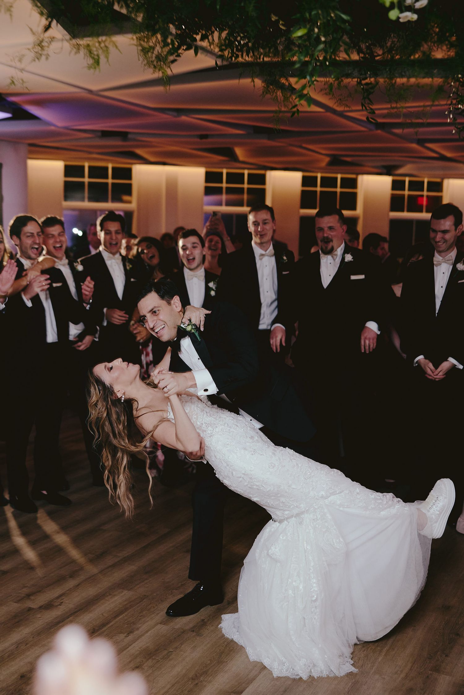 A couple performs a romantic dip dance move during their wedding reception as guests in formalwear look on and cheer.