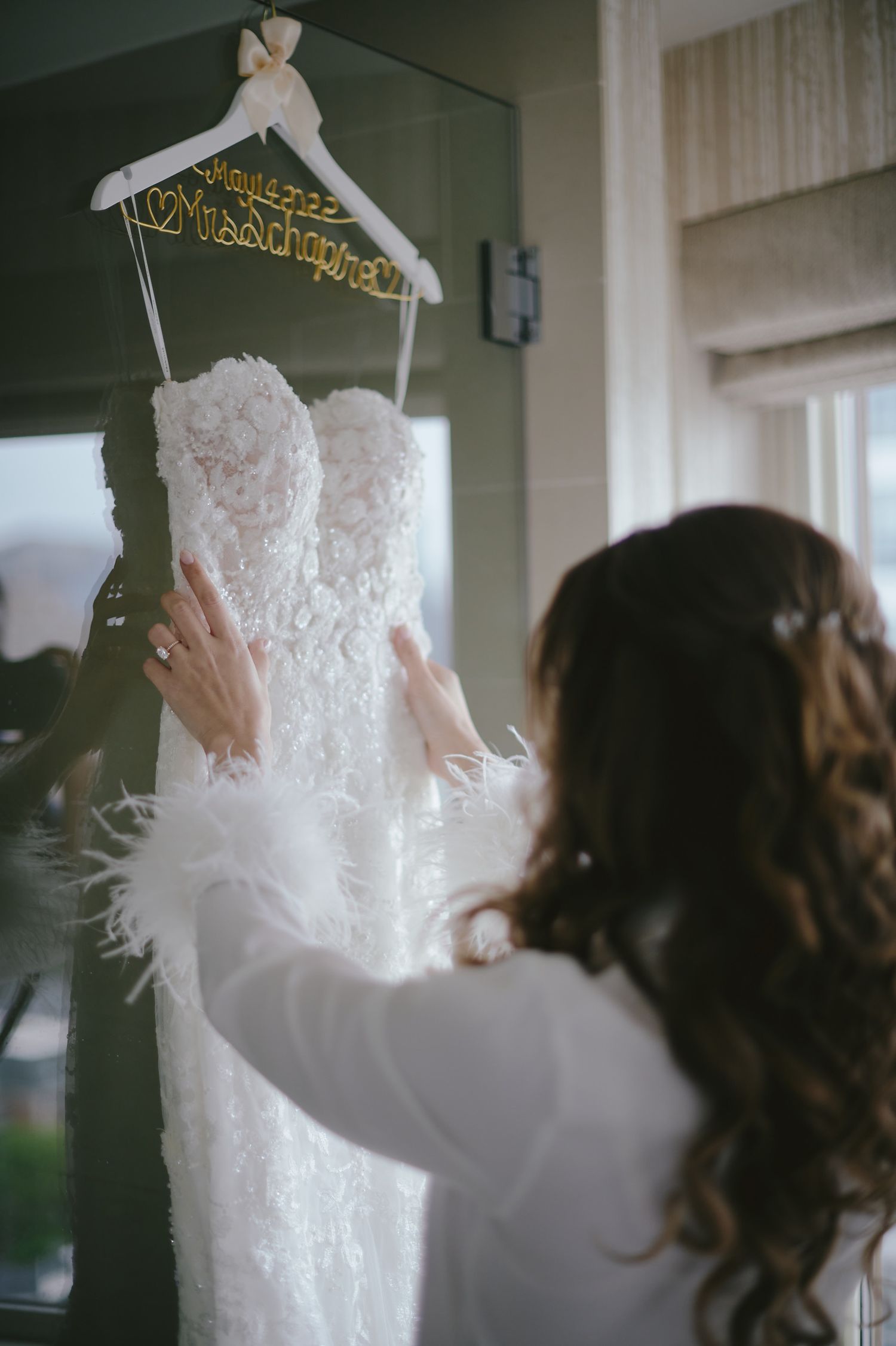 A white lace wedding dress hangs on a personalized wooden hanger as someone reaches up to touch the delicate fabric.