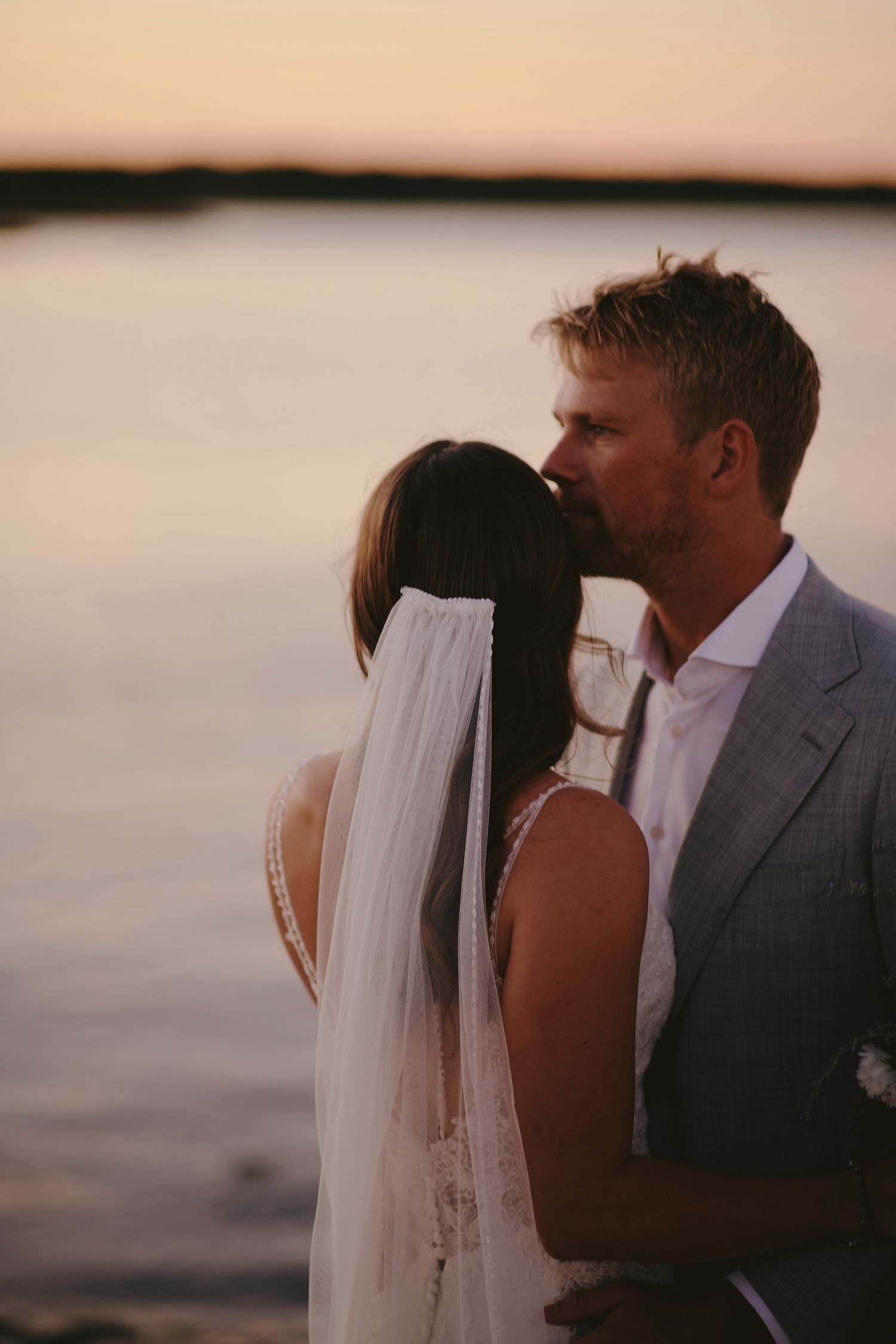A romantic sunset moment between newlyweds by the water with the bride wearing a flowing veil.