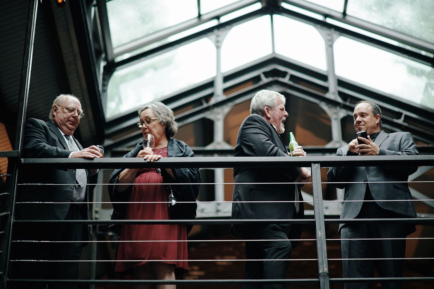 Group of older people in formal attire engaged in conversation while standing on a modern balcony with glass ceiling.
