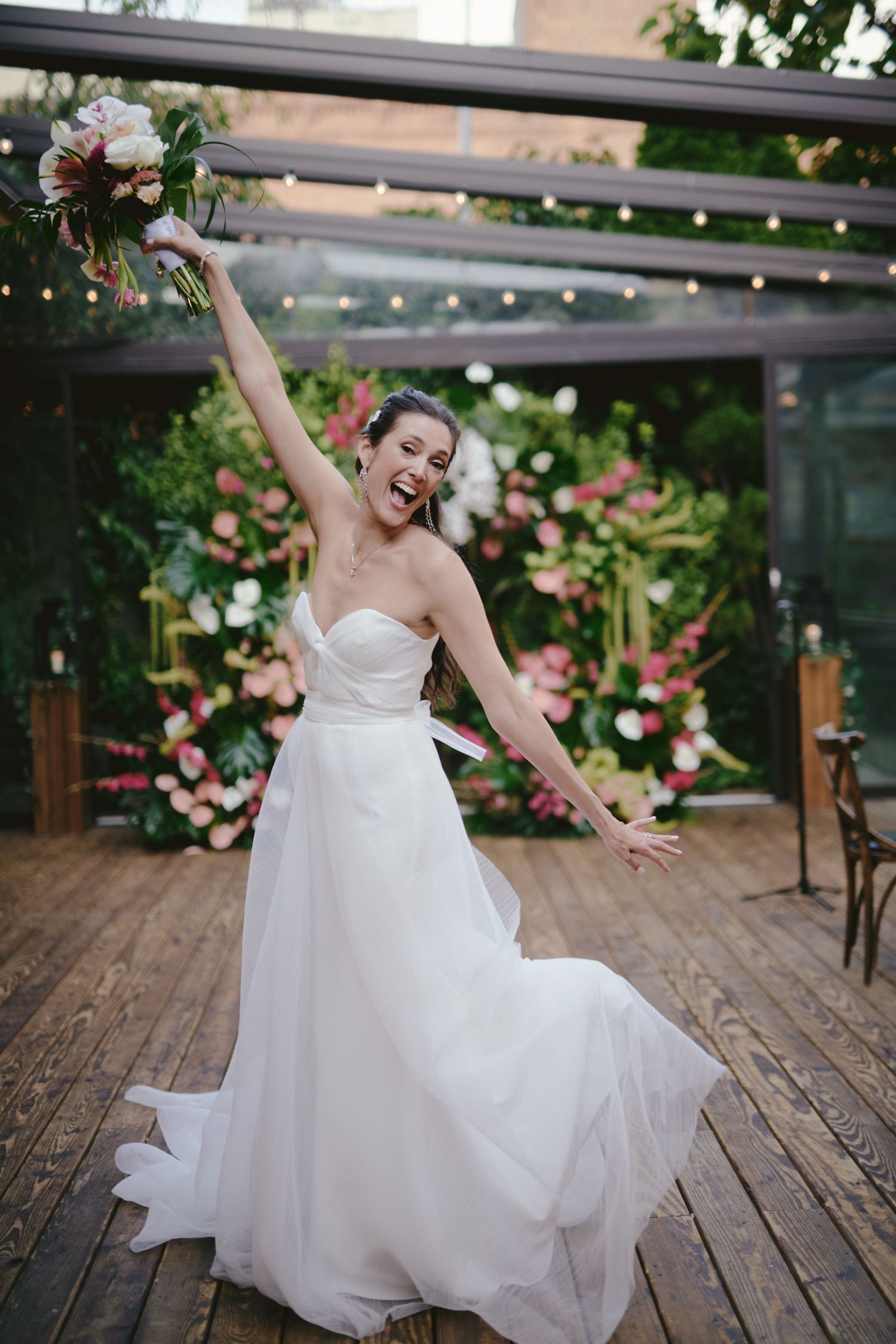 Joyful bride in strapless white wedding dress dances on wooden deck surrounded by pink floral decorations.