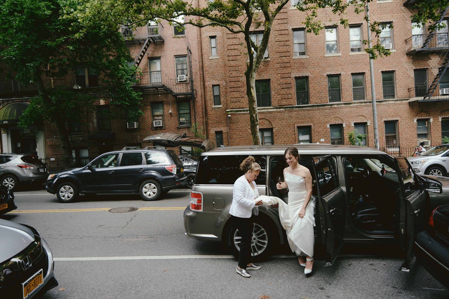 A couple arrives in formal dress beside a car on a tree-lined city street with brick apartment buildings.