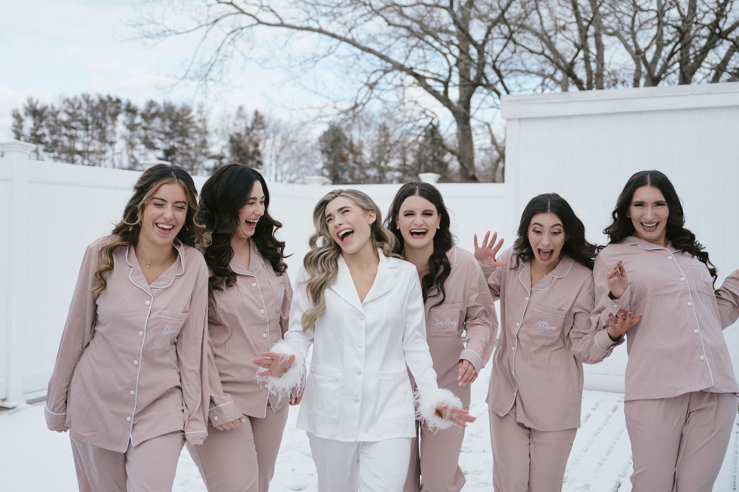 A bride in white pajamas laughs with five bridesmaids in pink matching sets outdoors in a snowy winter setting.