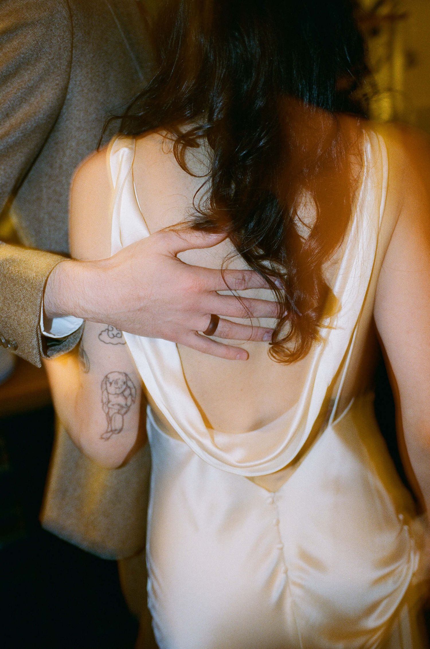 Close-up view of a person in a white satin dress with dark hair being embraced, showing a small tattoo on their arm.