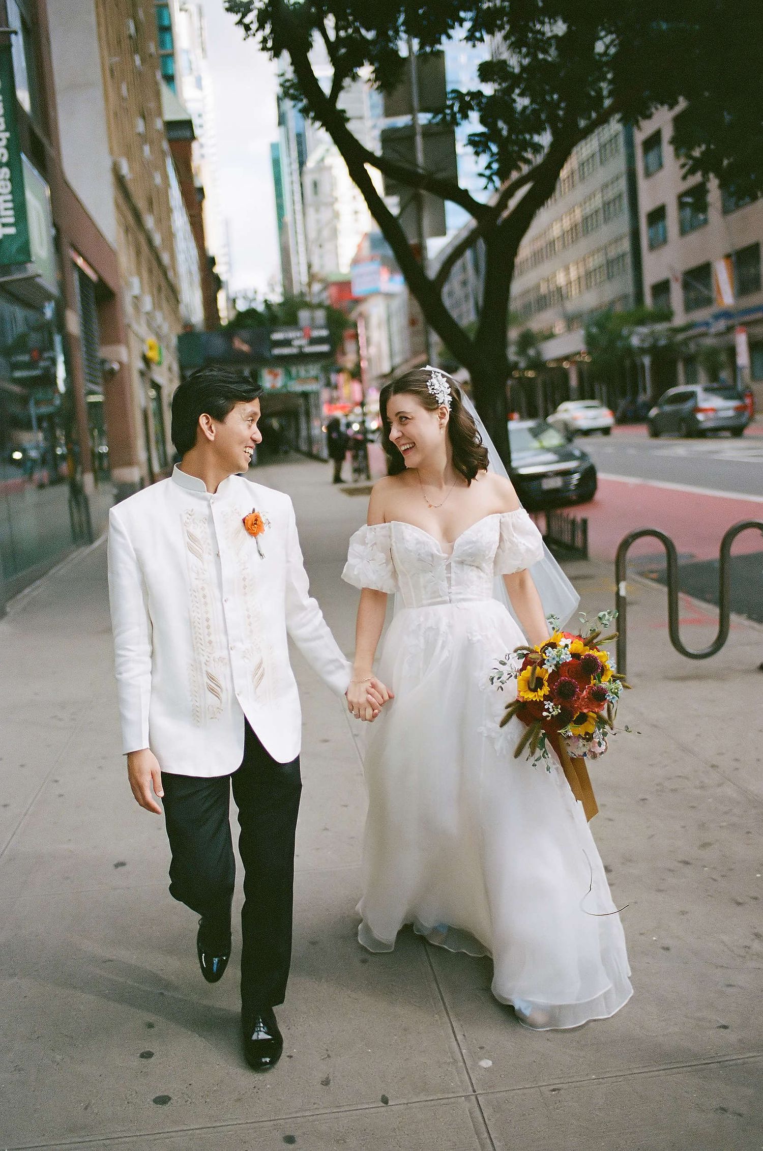 Newlyweds walking down a city street as bride holds sunflower bouquet and groom wears white jacket with orange boutonniere.