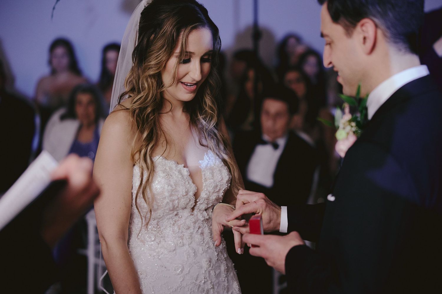Exchange of wedding rings during romantic indoor ceremony with bride in lace plunging neckline dress.