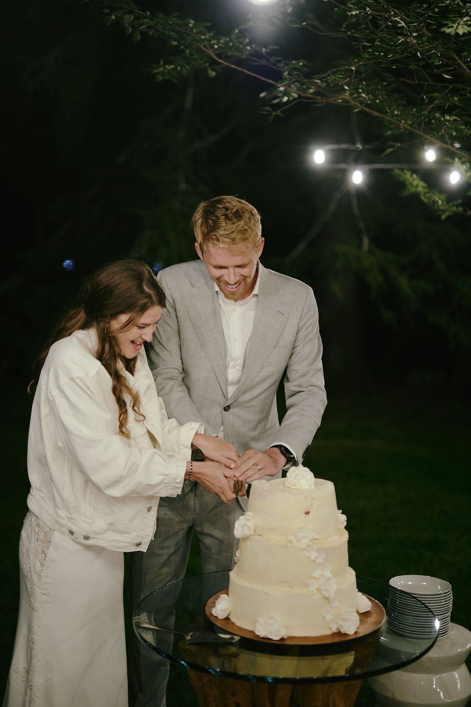 A couple cuts their elegant white wedding cake together at an outdoor evening reception under string lights.