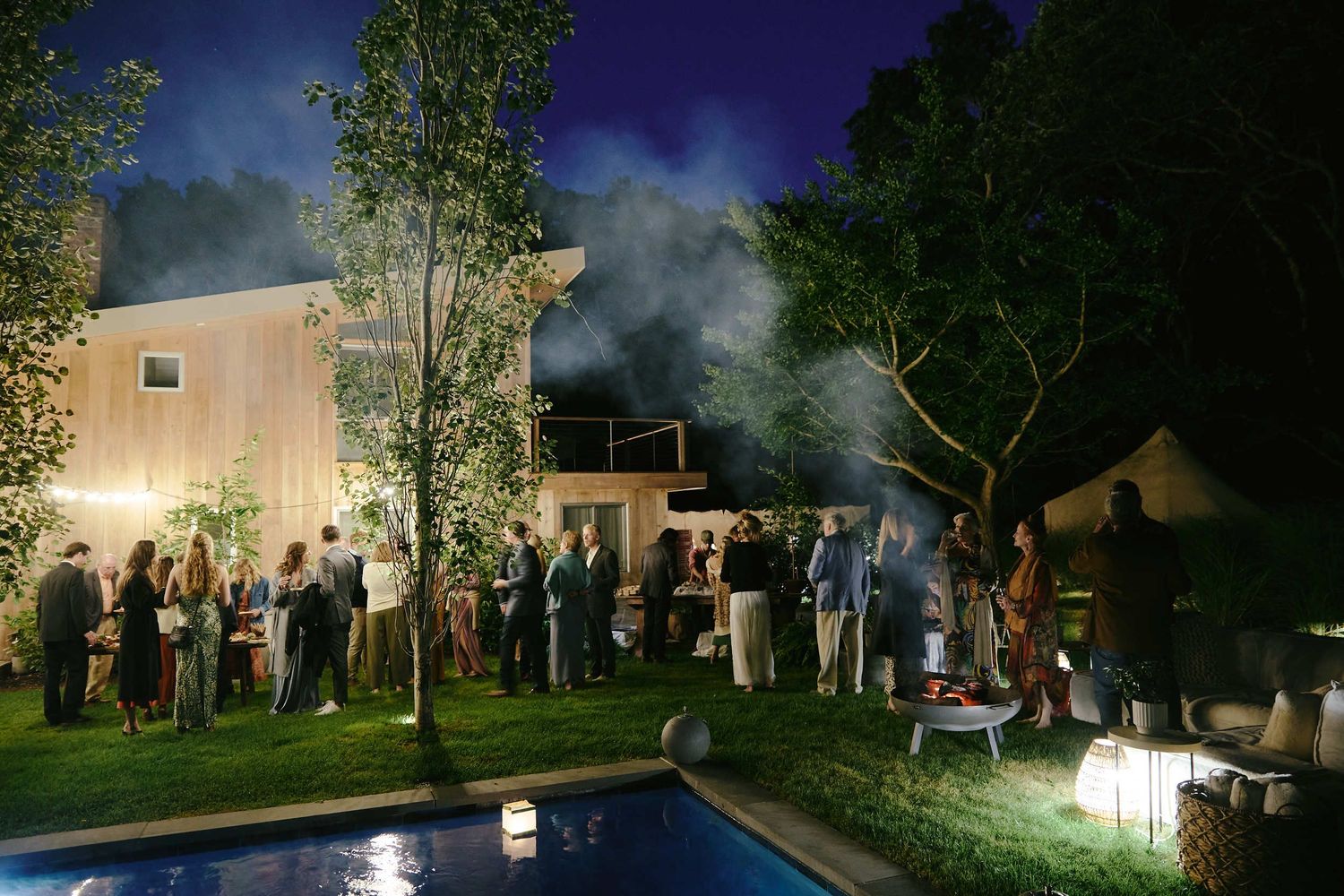Party guests gather around a swimming pool at night with ambient lighting and smoke effects creating a moody atmosphere.