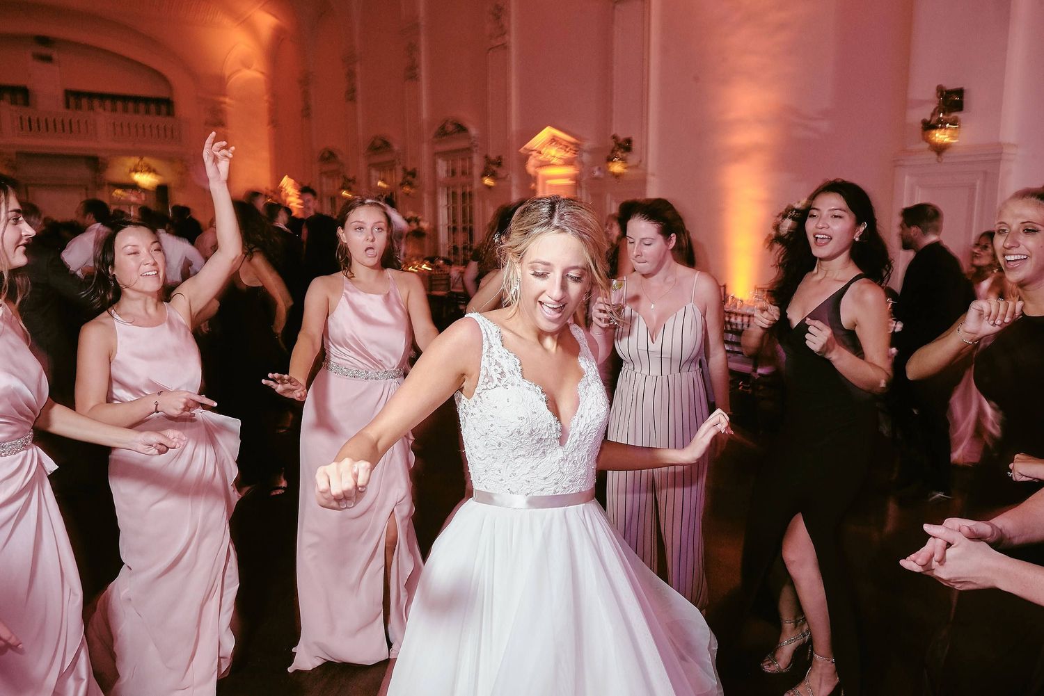 Wedding guests dance together in an elegant reception venue with pink lighting and a crowd on the dance floor.
