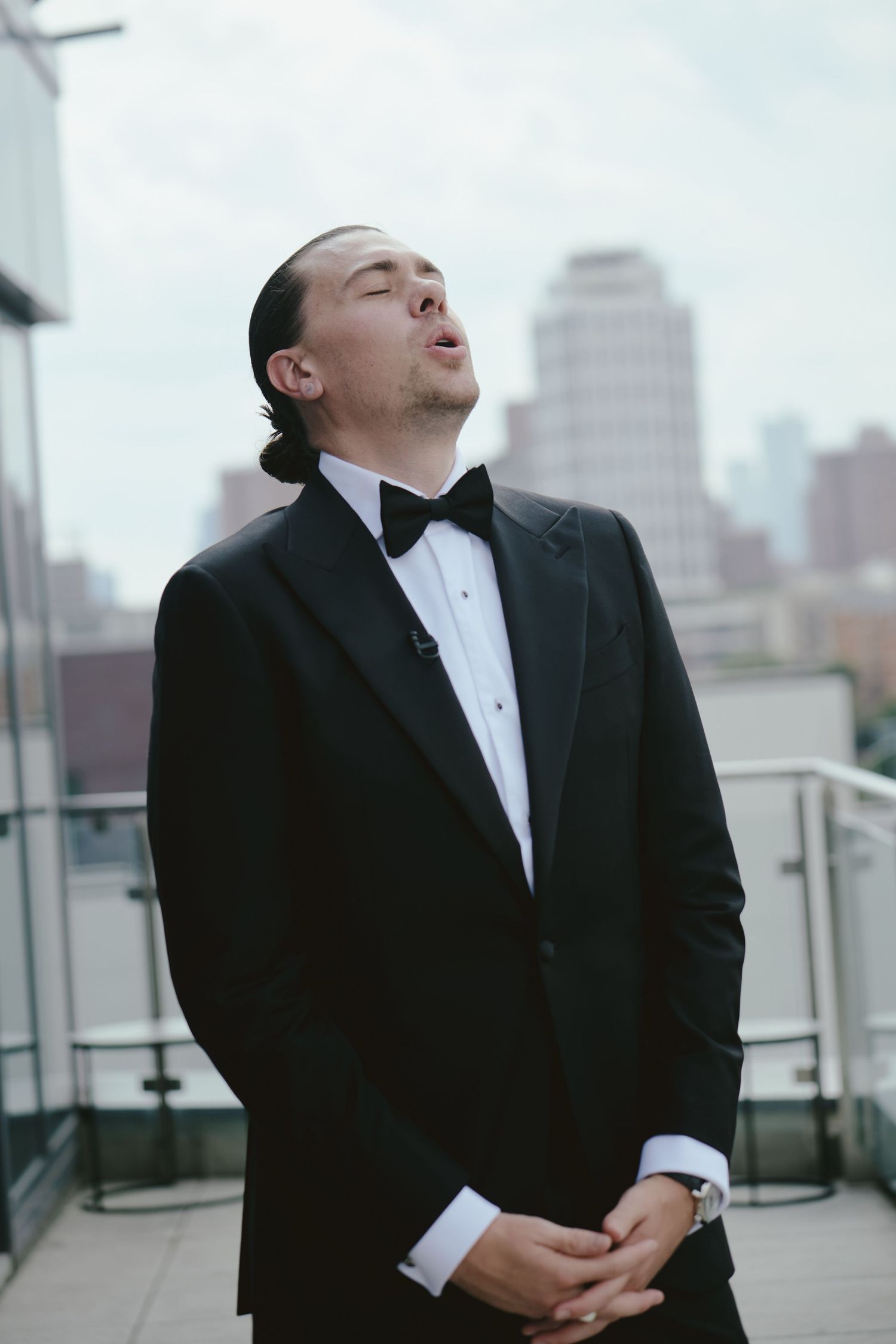 A person in a tuxedo and bow tie tilts their head back while standing on a rooftop with city buildings behind them.