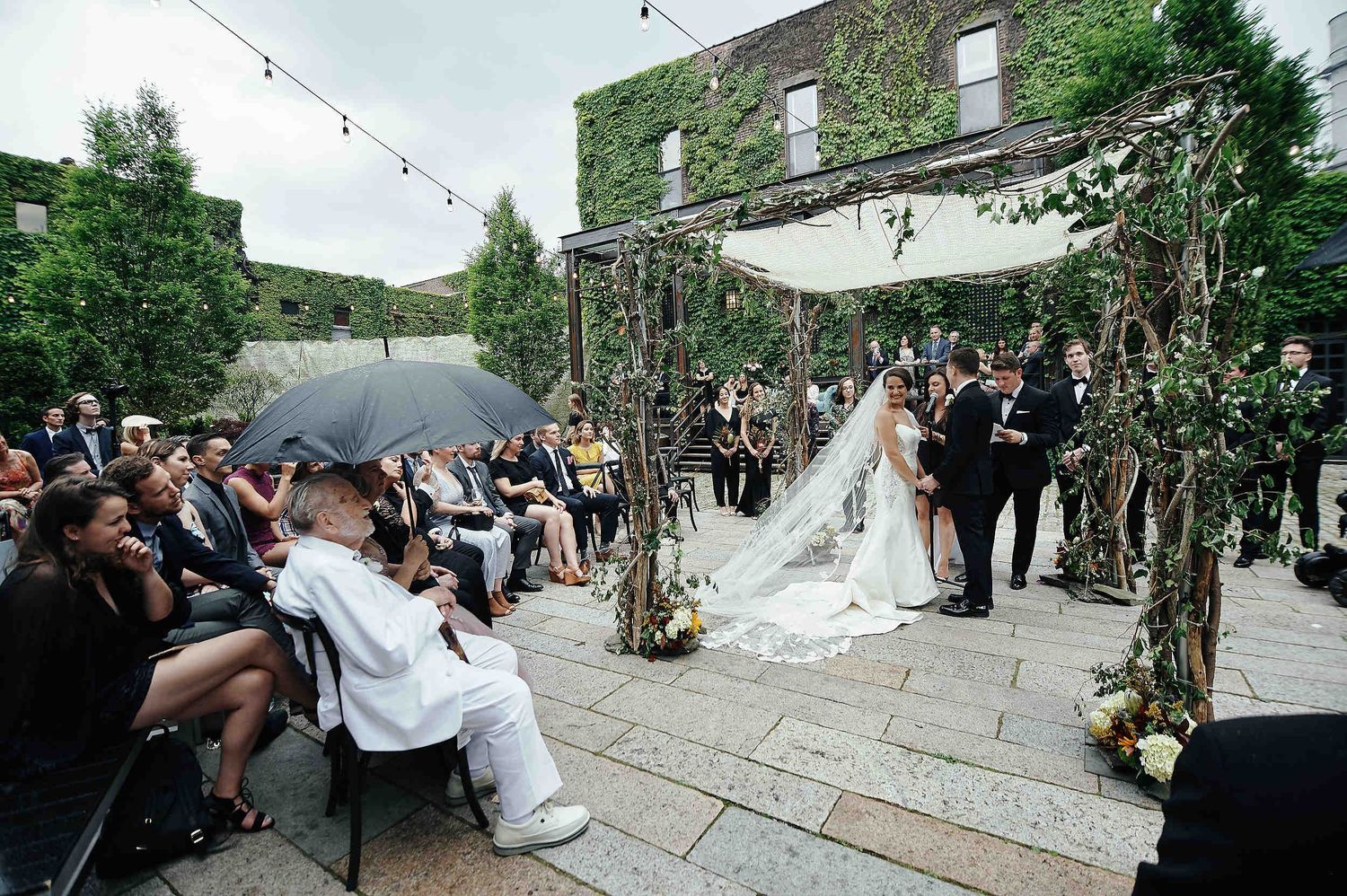 An outdoor wedding ceremony taking place on a stone patio with greenery-covered walls and string lights overhead.