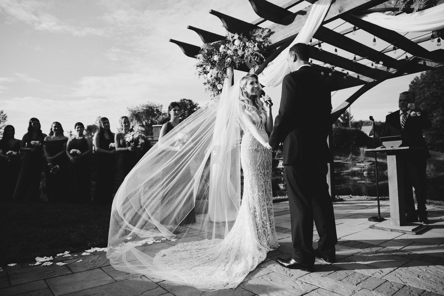 A black and white wedding photo capturing a bride in a flowing veil during an outdoor ceremony under a wooden pergola.