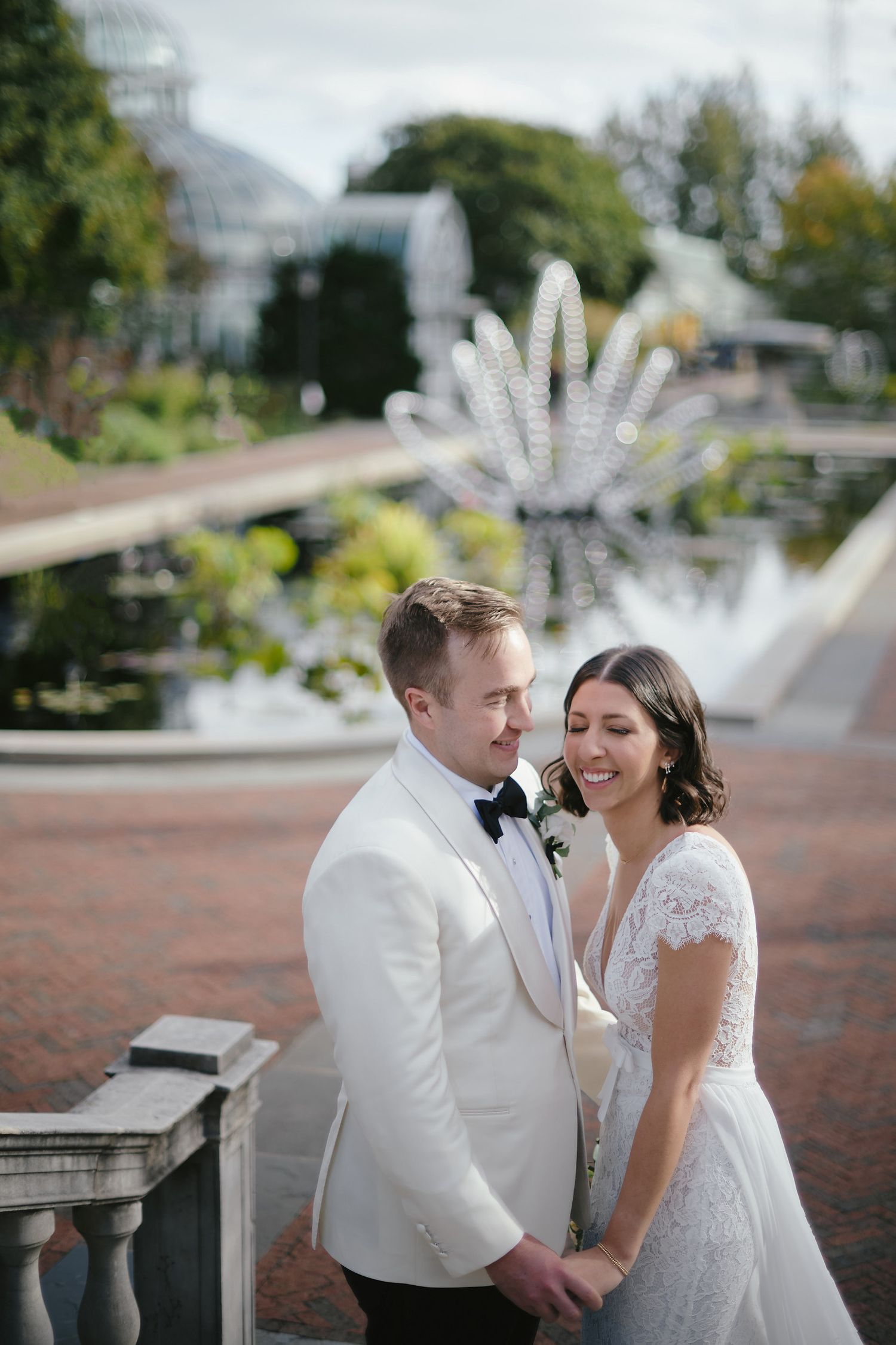 A couple in wedding attire share an intimate moment by a garden fountain with a decorative light feature in the background.