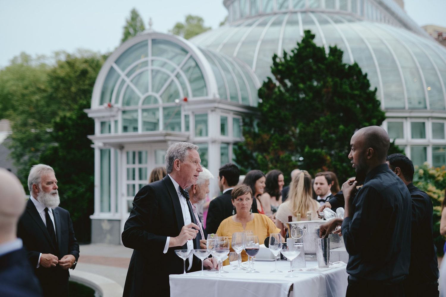 Garden party guests mingle during an elegant reception beneath a Victorian-style glass conservatory.