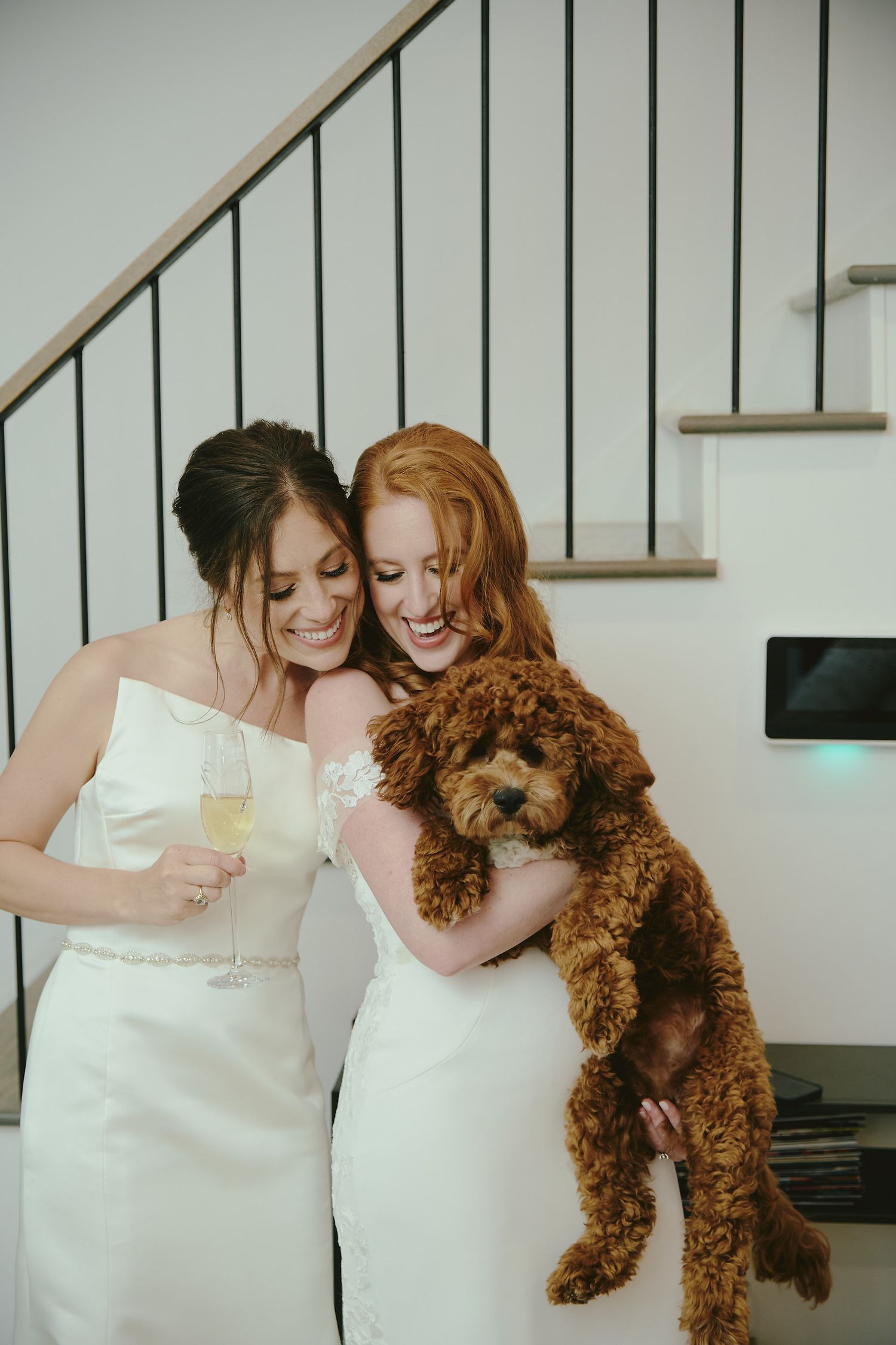 Two brides in white dresses share a joyful moment with their adorable goldendoodle puppy during their wedding celebration.