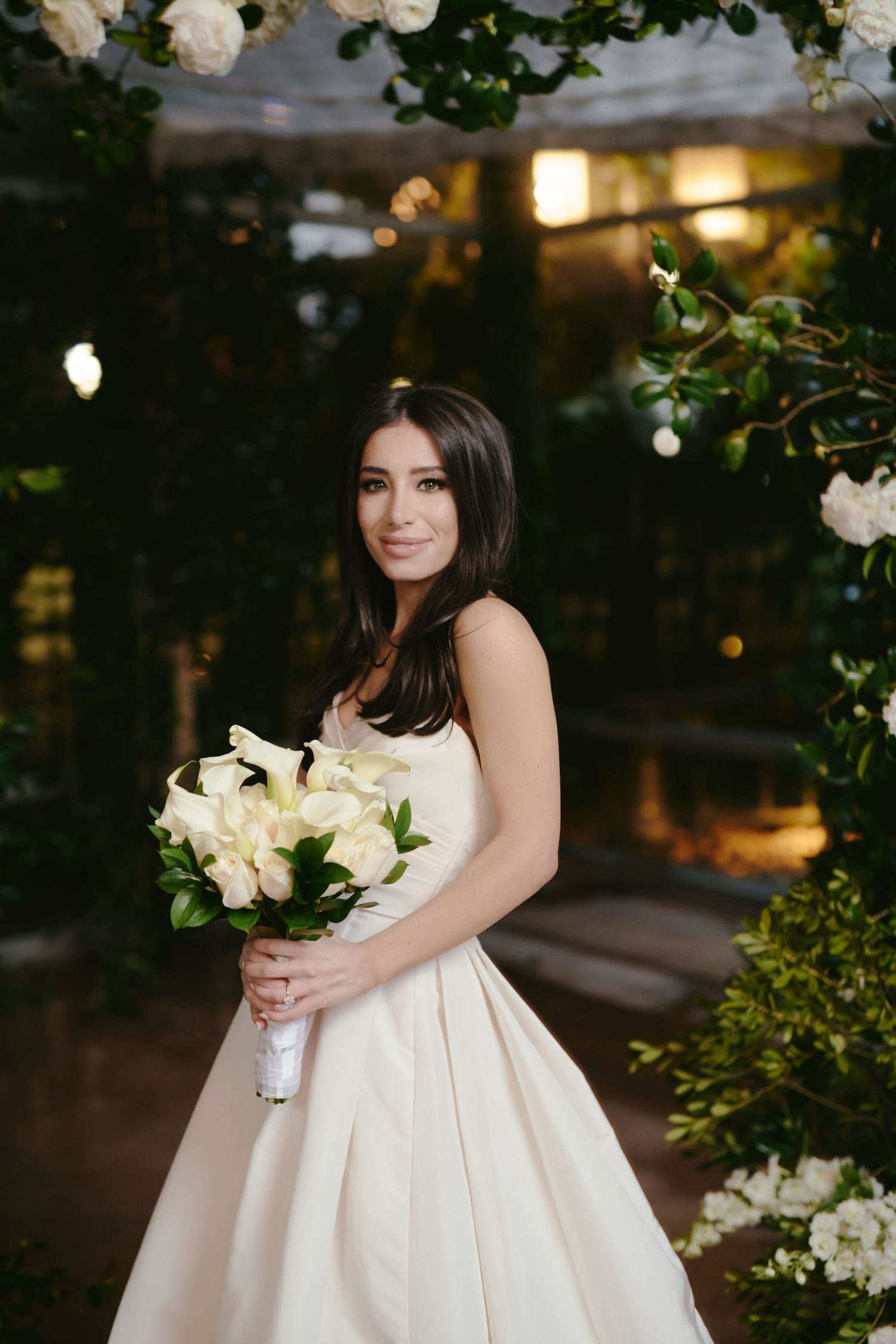 A bride in a classic white ball gown holds a bouquet of white lilies in a garden at sunset.