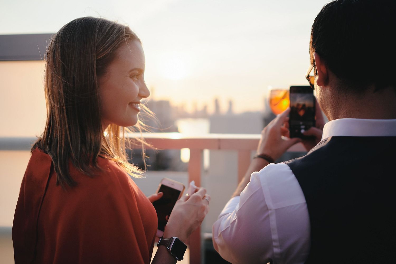 A couple enjoying sunset drinks while taking photos on a rooftop terrace.