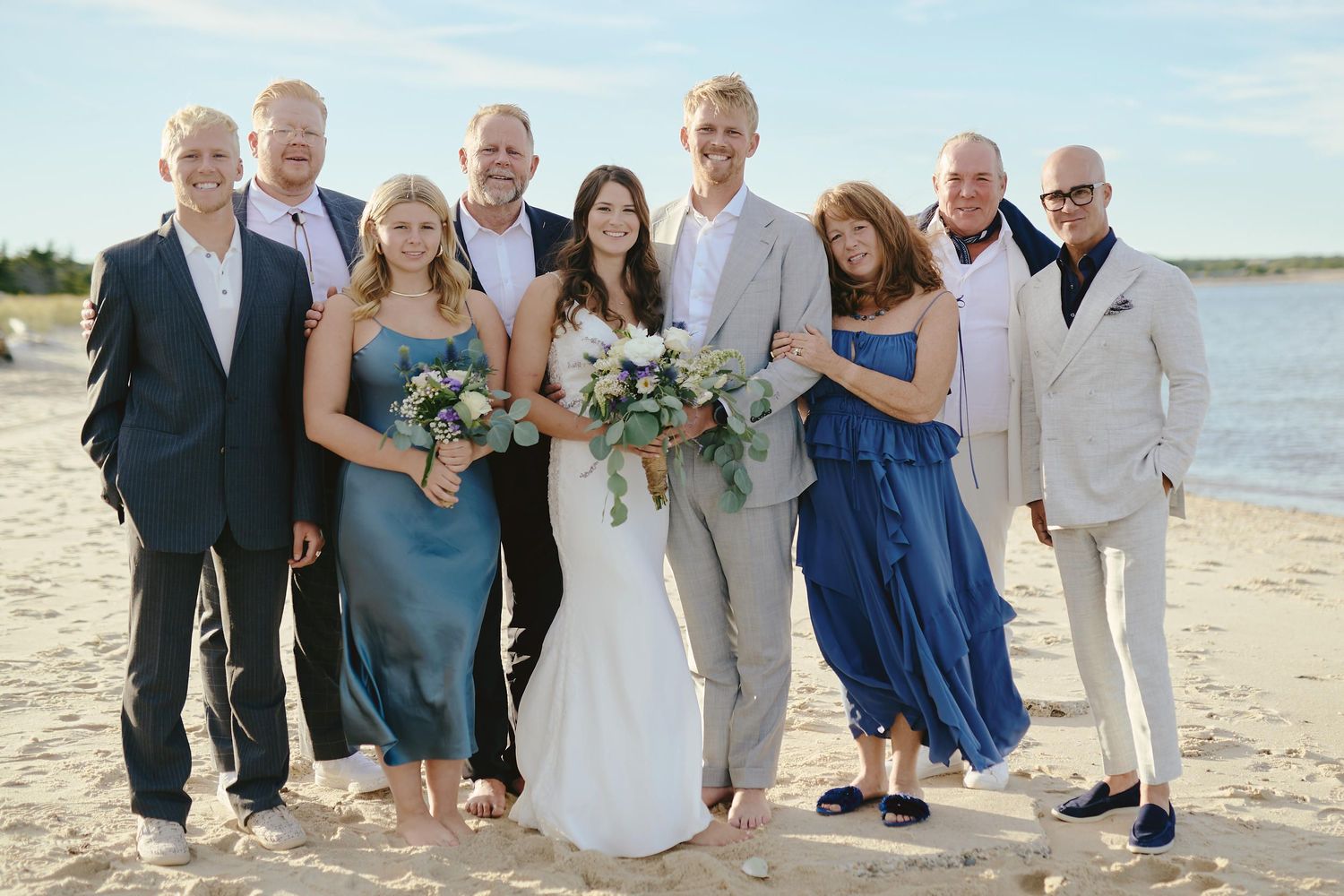 A wedding party wearing formal attire poses together on a sunny beach with the ocean in the background.