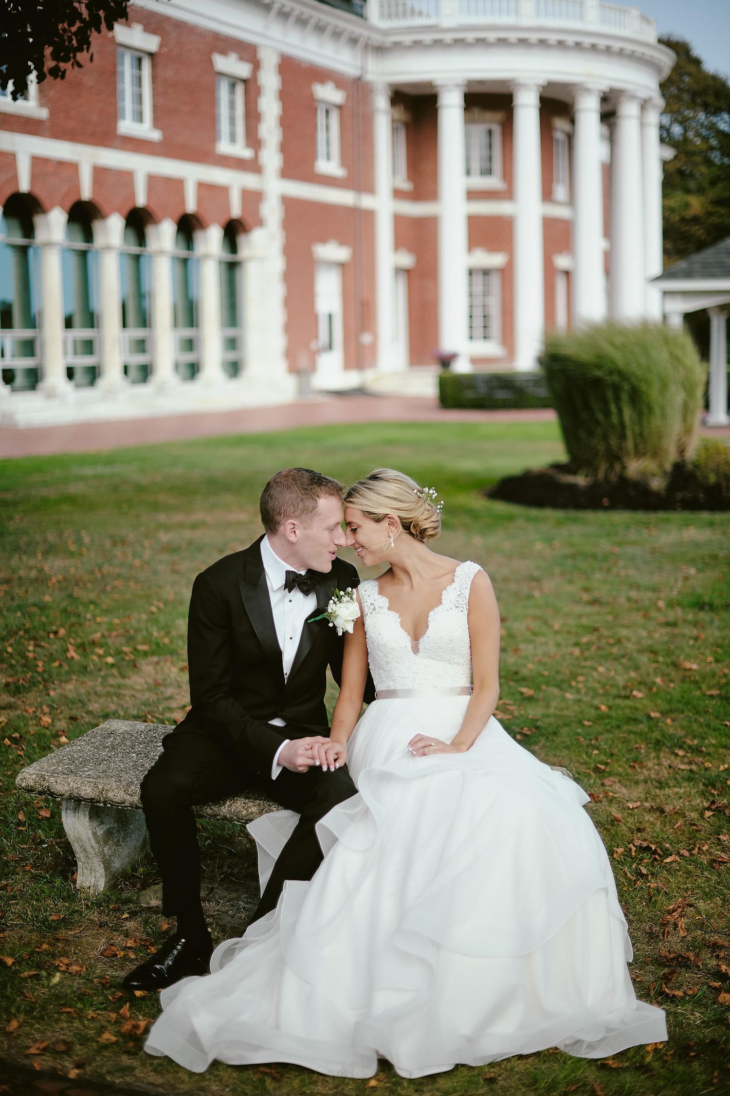 Newlyweds share intimate moment on stone bench outside elegant mansion with white columns and pink brick facade.