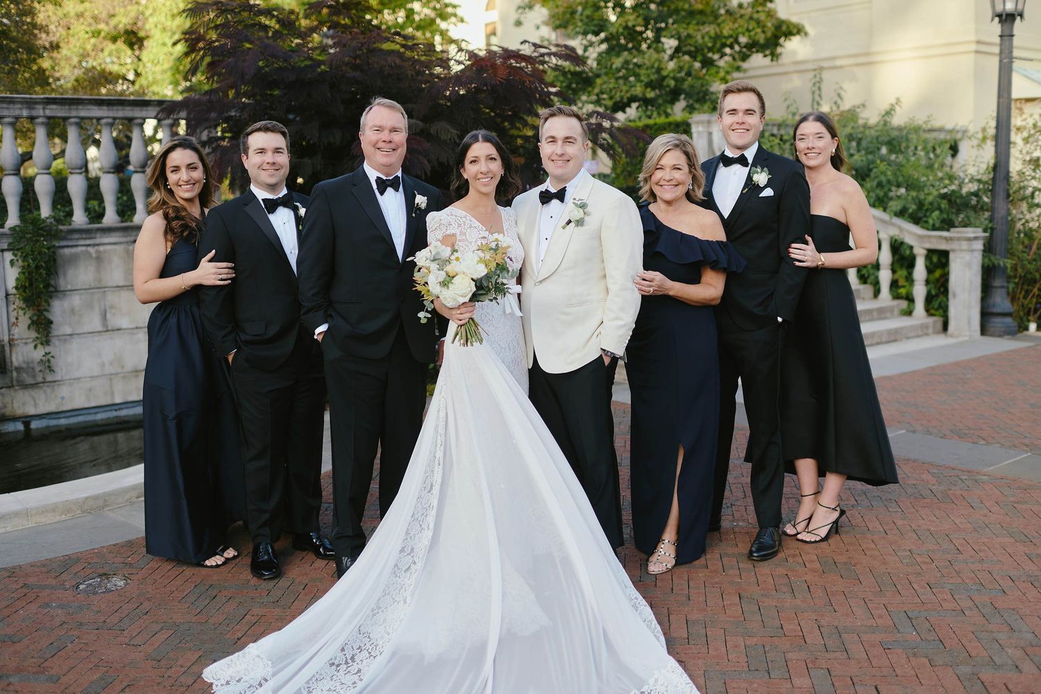 A wedding party in formal attire poses together on brick pavement with the bride's train extending behind them.