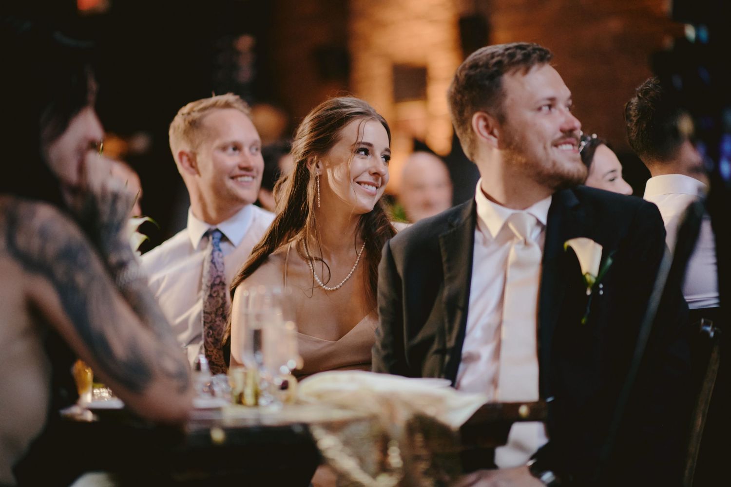 Wedding guests smile and laugh while seated at a reception table during an evening celebration.