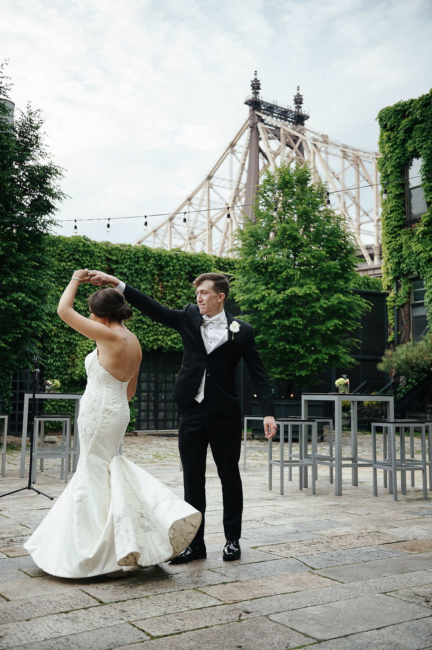 A wedding couple dancing together on a paved terrace with the Queensboro Bridge visible in the background.