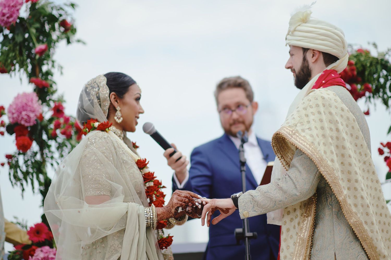 Couple exchanging rings during an outdoor Indian wedding ceremony with pink floral decorations.