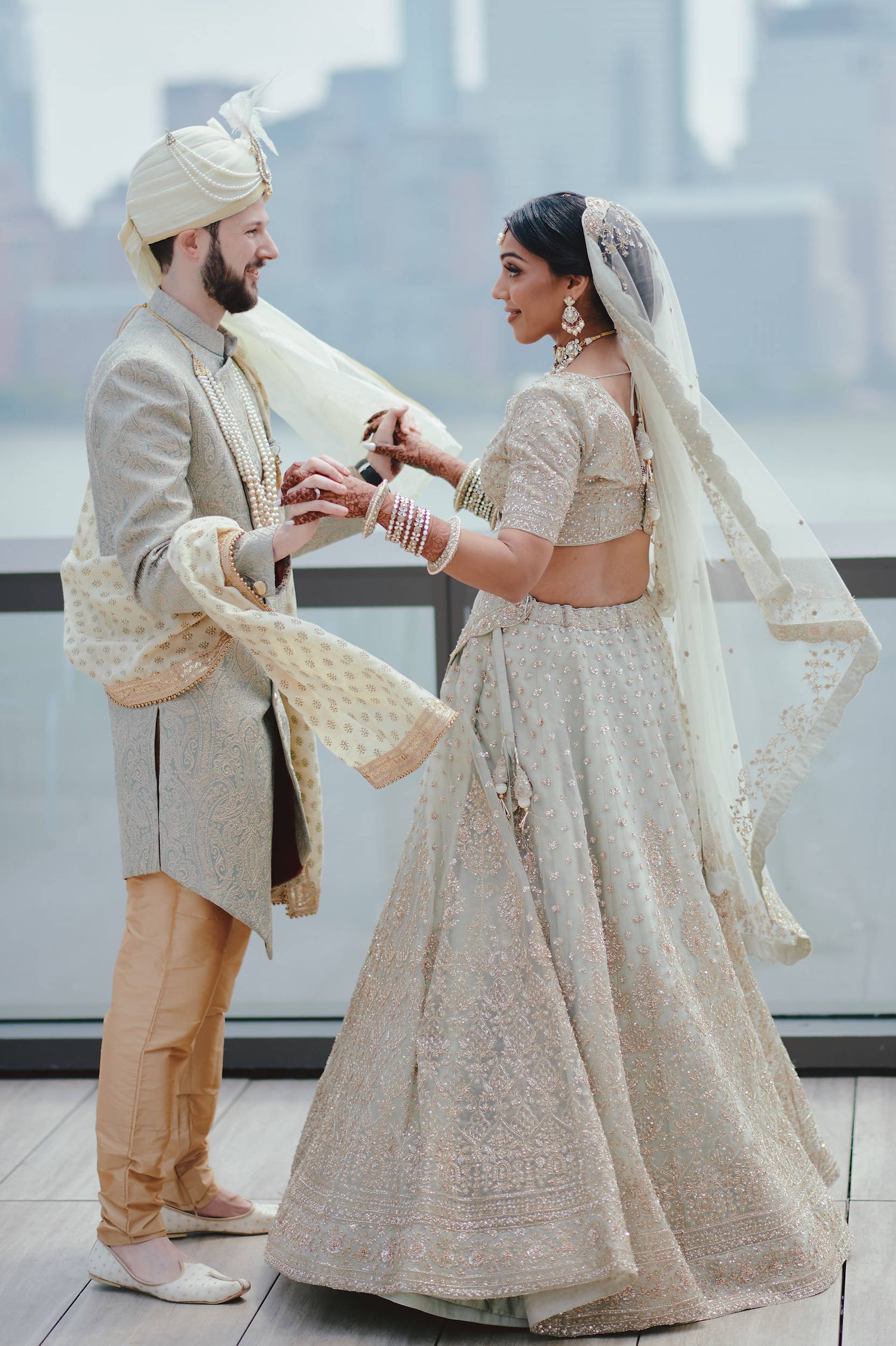 Newlyweds in elegant Indian wedding attire exchange rings on a rooftop terrace overlooking a city skyline.