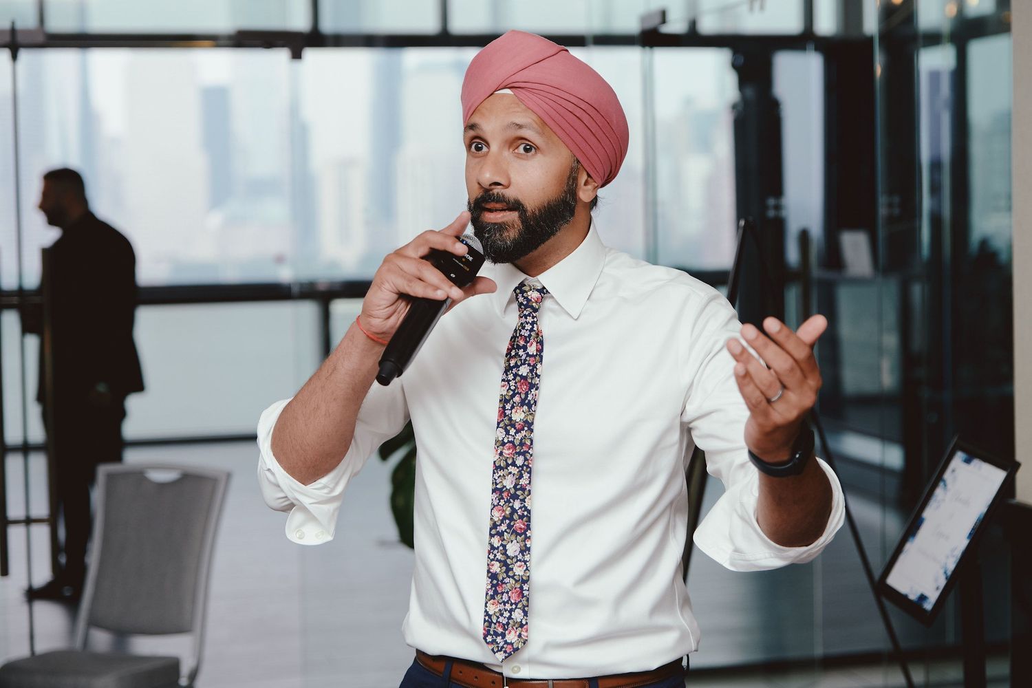 Business professional in white shirt and floral tie speaks gesturing with hands in modern glass office setting.