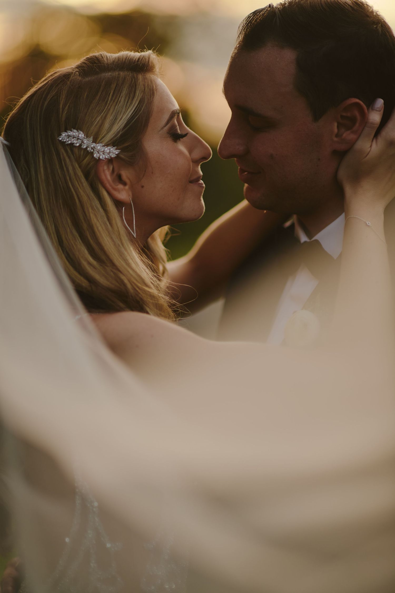 Romantic sunset wedding portrait showing close up of couple embracing through soft veil in golden light.