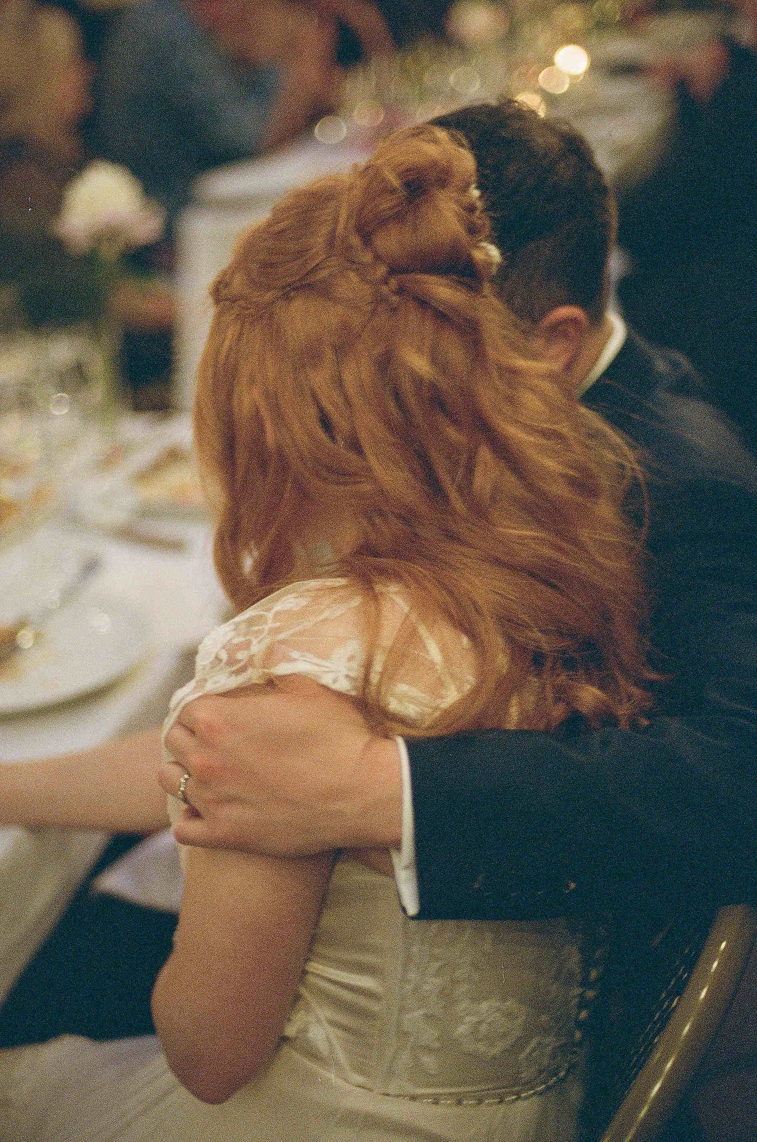 A couple embraces at a formal dinner event, with the womans flowing red hair and white dress visible from behind.