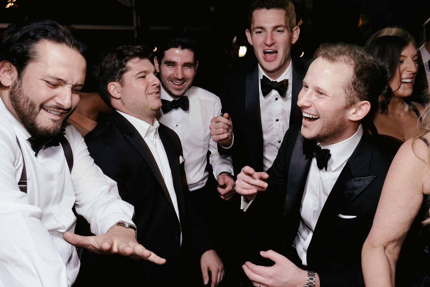 A group in formal black and white attire laugh and gesture animatedly at an elegant evening event.