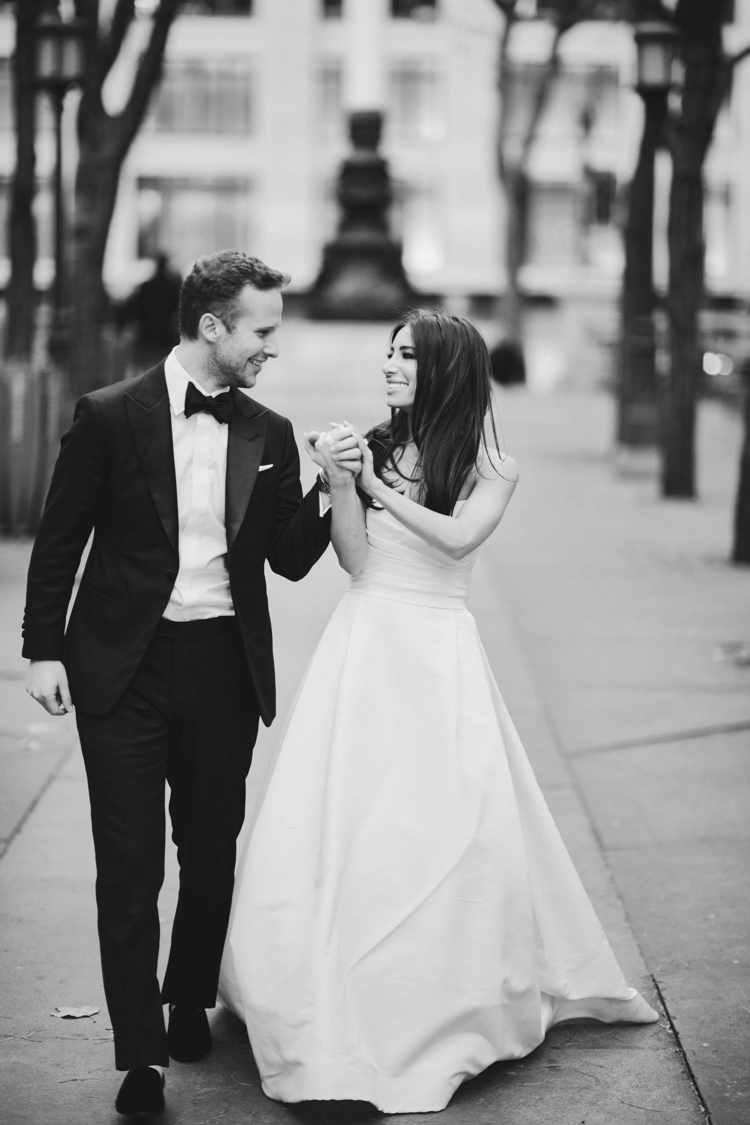 A bride in a white ballgown and groom in classic tuxedo walk together on a city sidewalk in black and white photography.