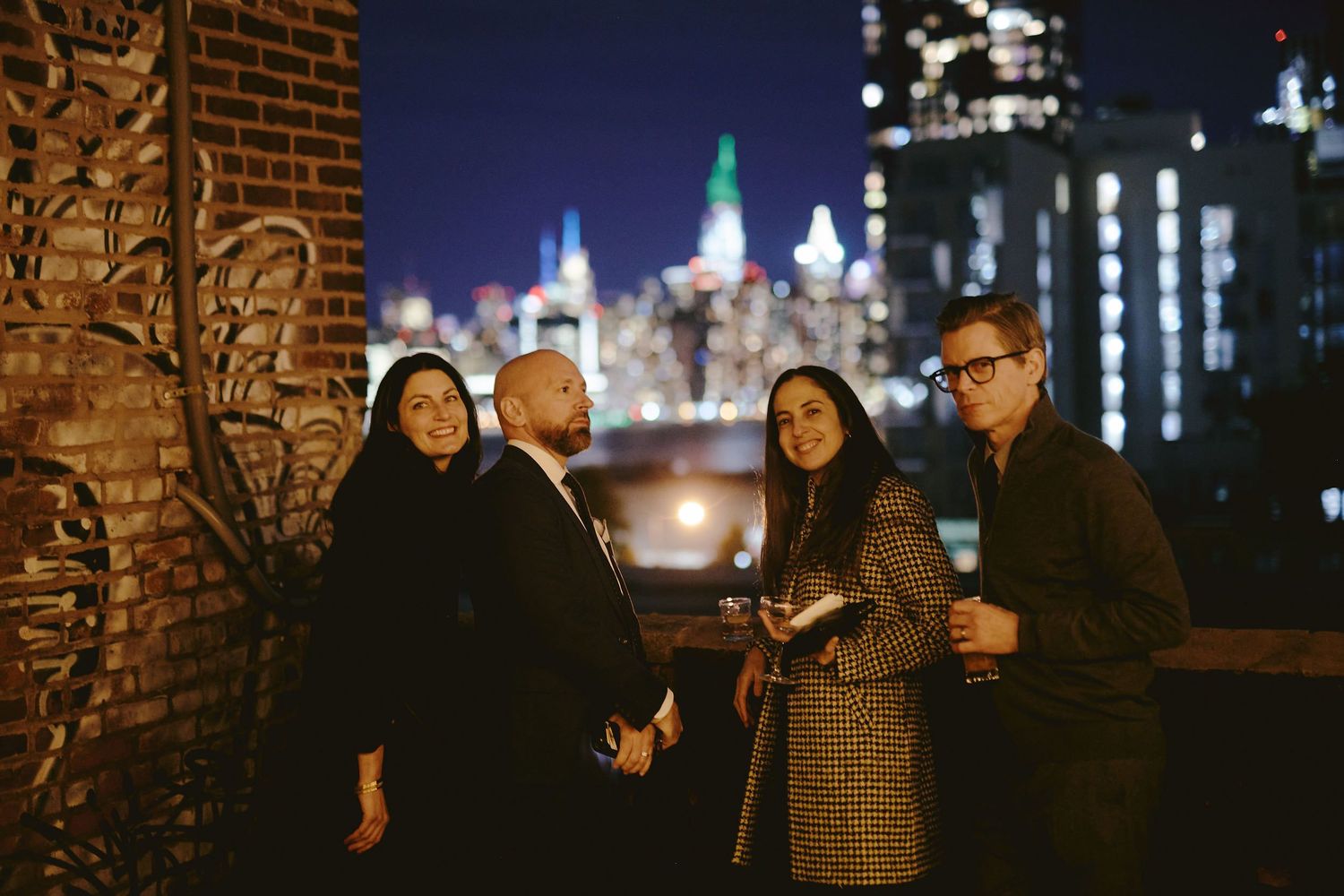 Two couples stand together at night with illuminated city buildings behind them glowing in the distance.