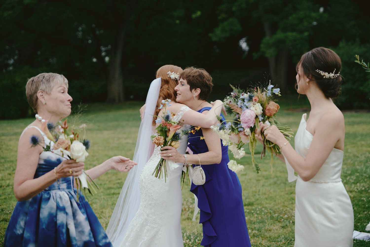 Wedding party members share laughter and joyful moments while holding floral bouquets on a grassy lawn.