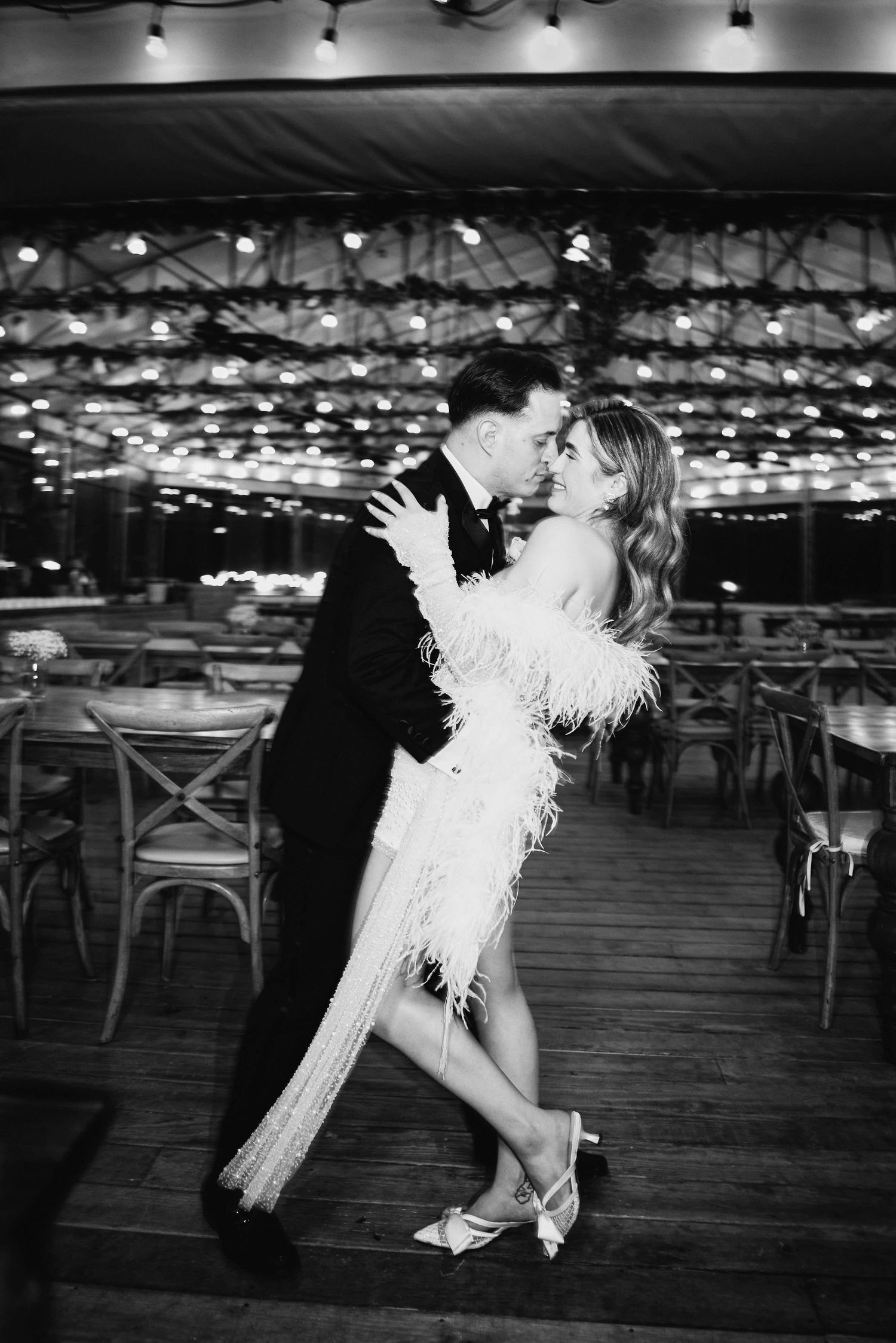 Couple sharing a romantic kiss while dancing in a dimly lit venue with string lights, man in tuxedo, woman in feathered dress.