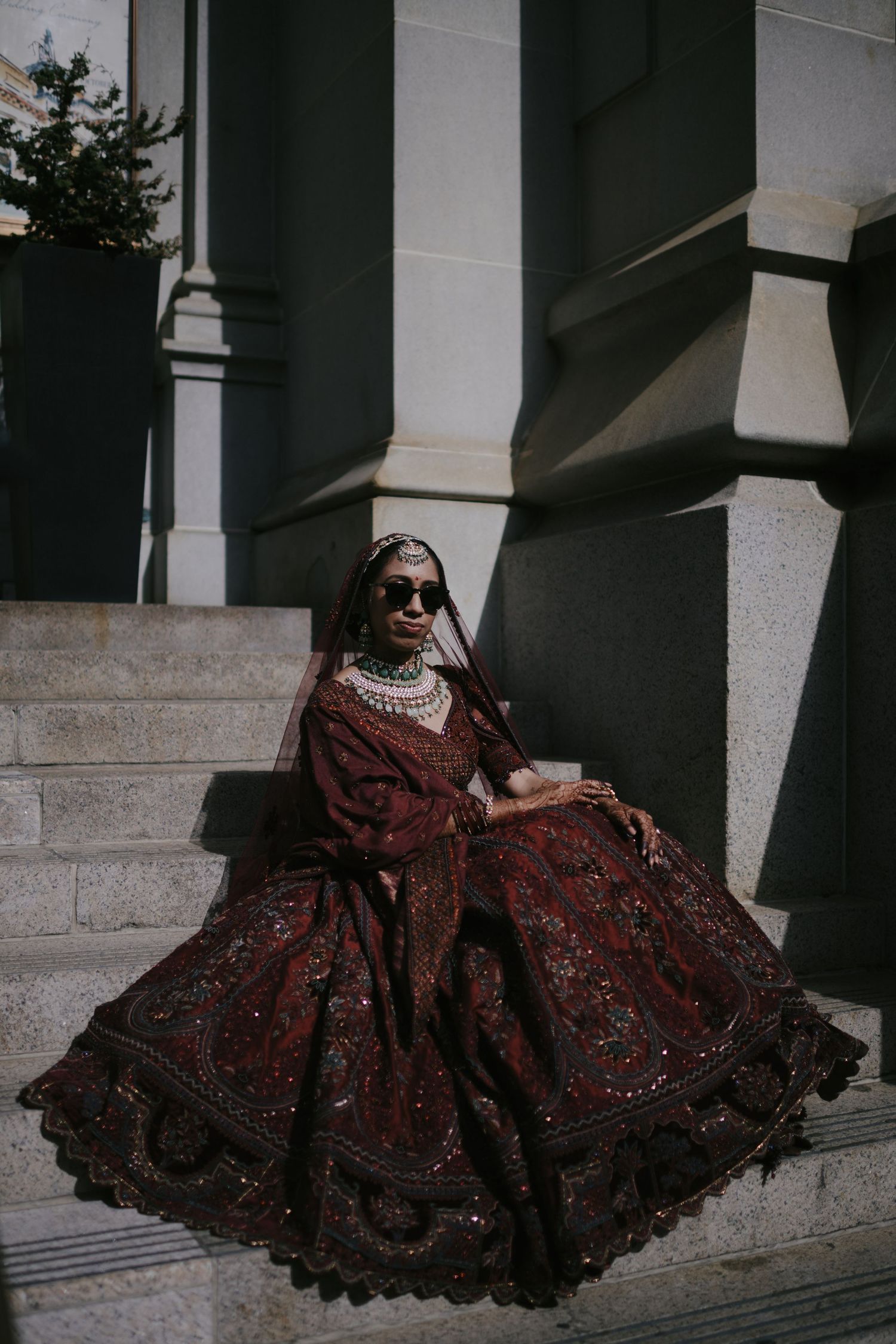 Woman in ornate dark red bridal lehenga with jewelry and sunglasses seated on stone steps.