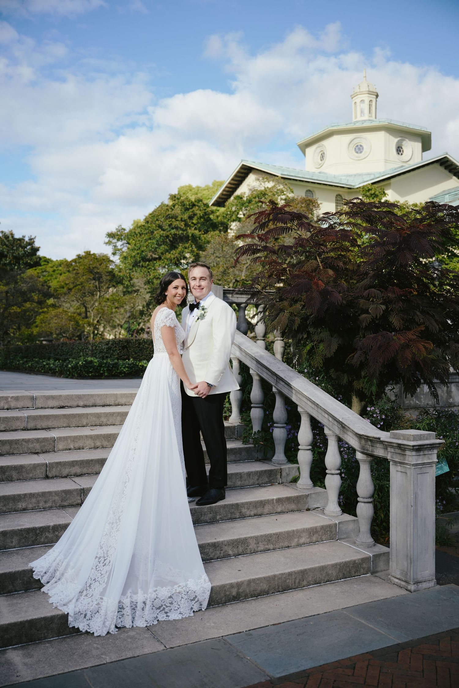 Bride and groom pose on elegant stone staircase with white balustrade outside a classic venue on their wedding day.