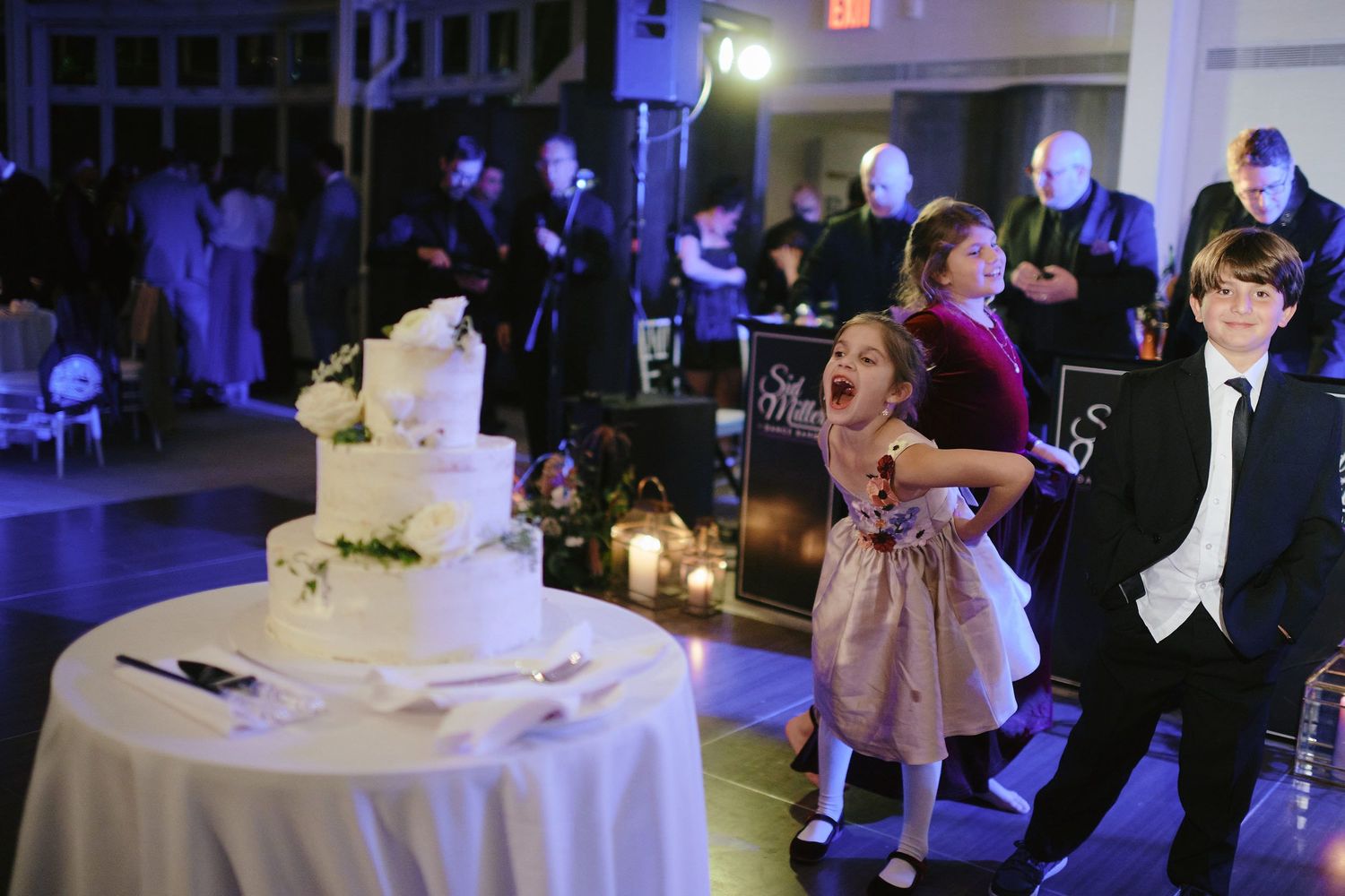 Wedding cake displayed on table while guests dance at dimly lit reception with purple lighting.